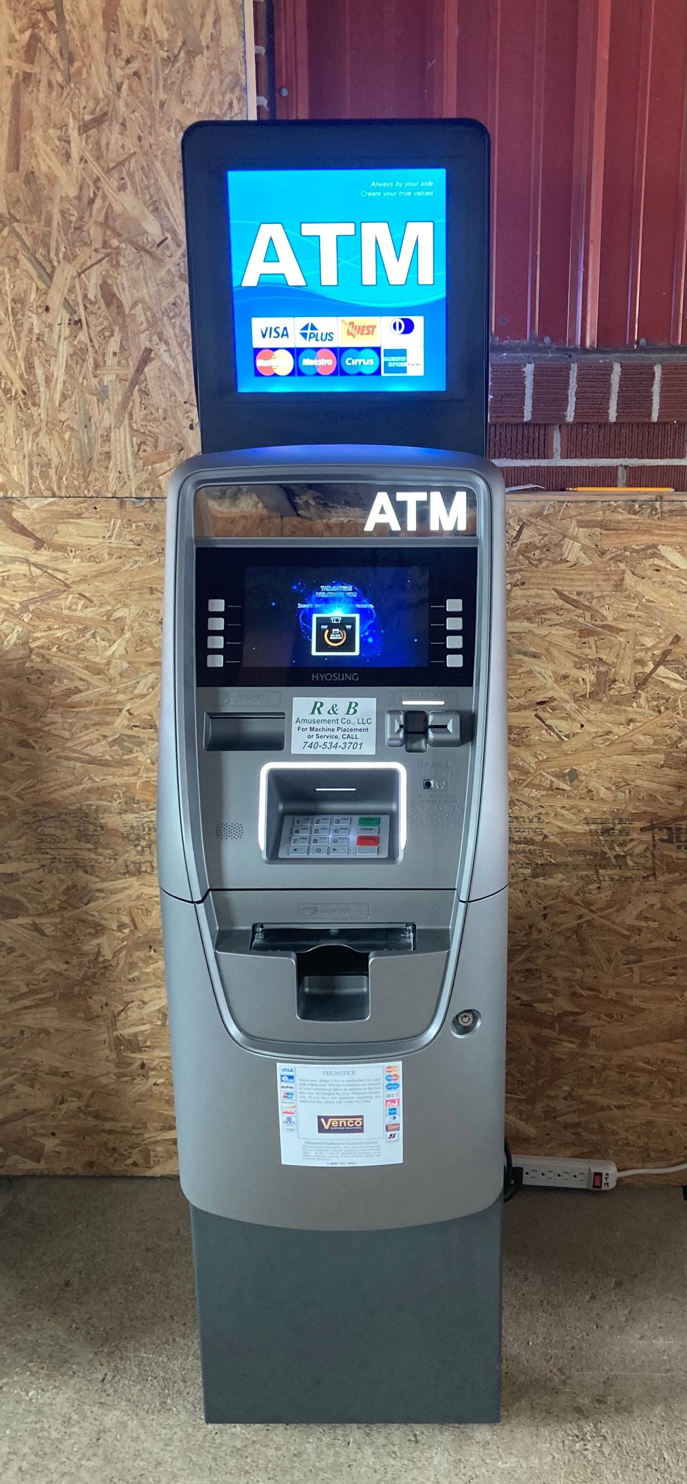A standalone ATM with a digital top screen and main display, standing in front of a light-colored wooden-textured wall.
