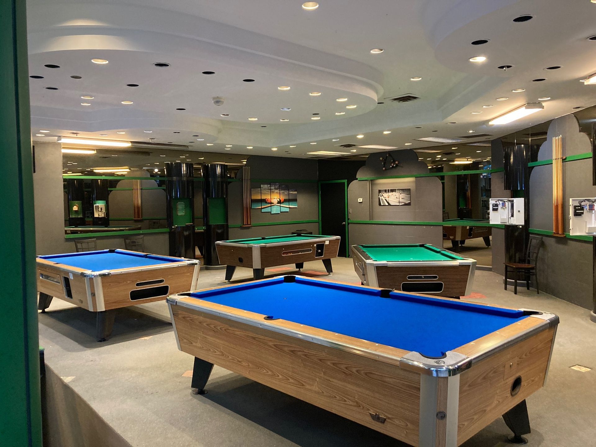 A room with multiple pool tables featuring blue and green felt surfaces, set against a backdrop of dark, paneled walls.