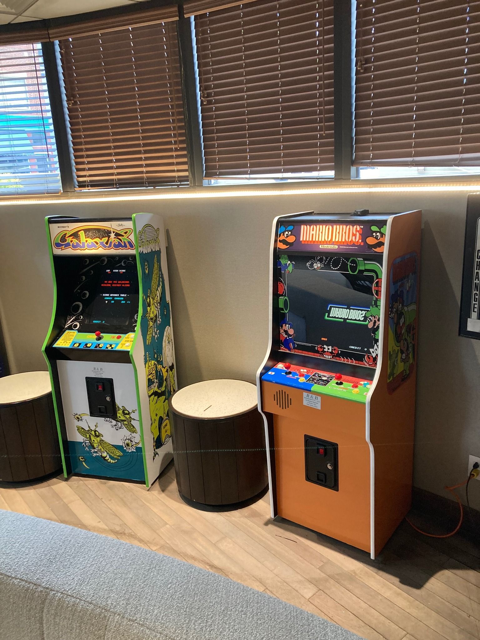 Two retro-style arcade cabinets stand against a beige wall by a window, separated by wooden stump-shaped stools.