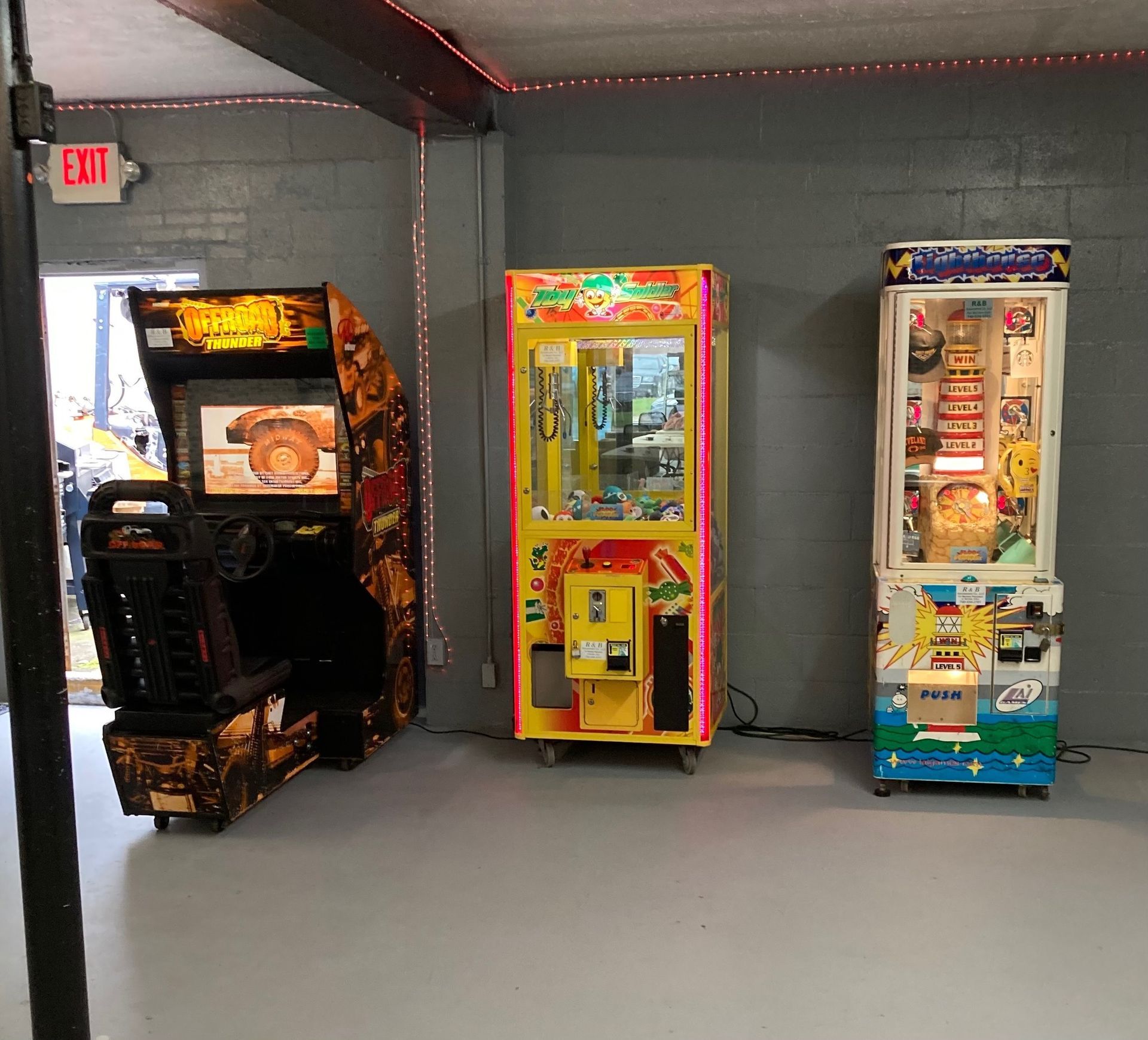 Three arcade game cabinets, including a racing game and two prize machines, stand against a grey wall in an indoor space.
