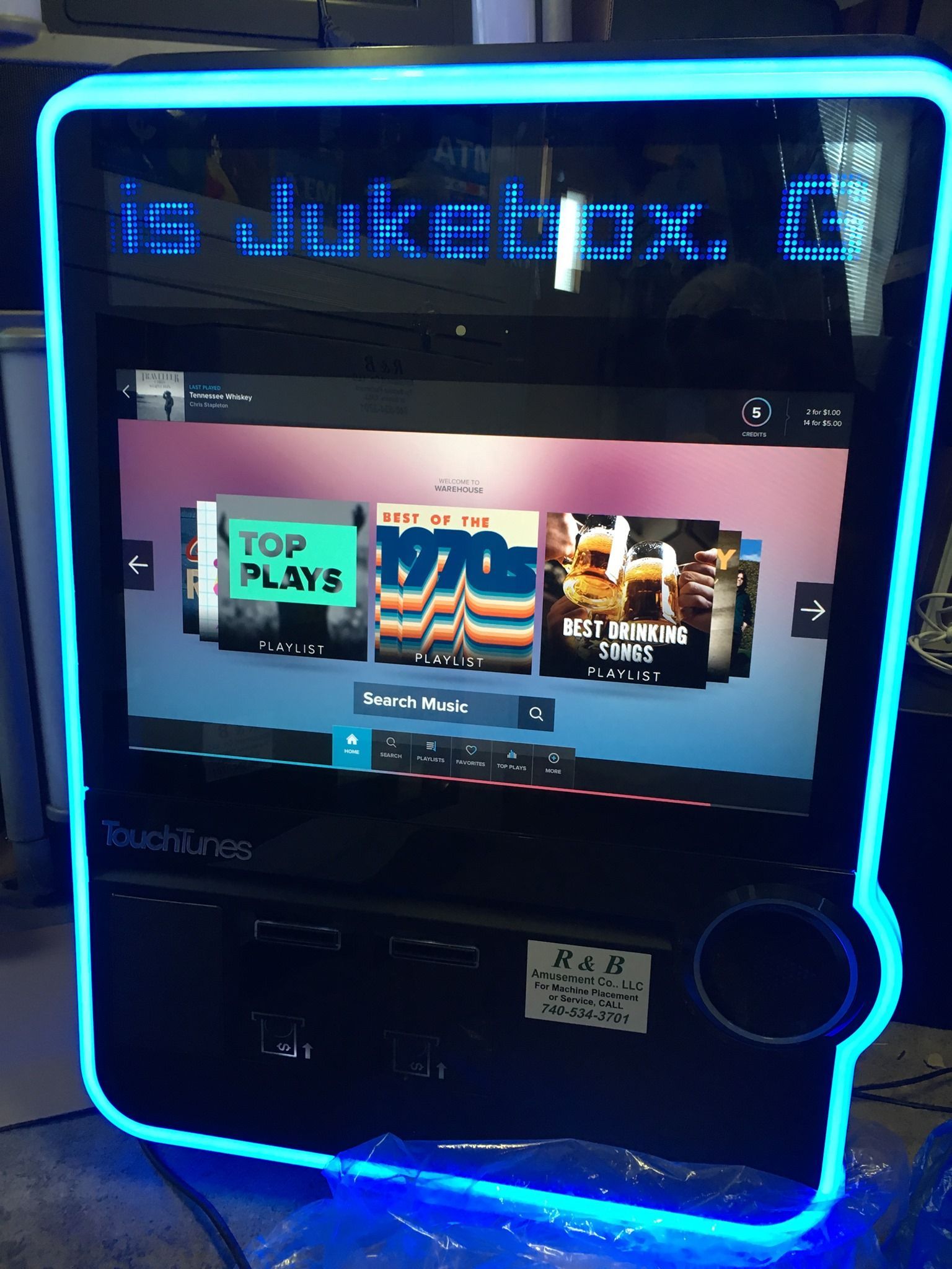 A TouchTunes digital jukebox with a bright neon blue frame, displaying a music selection screen with album covers.