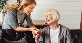 A woman is holding the hand of an elderly woman who is sitting on a couch.