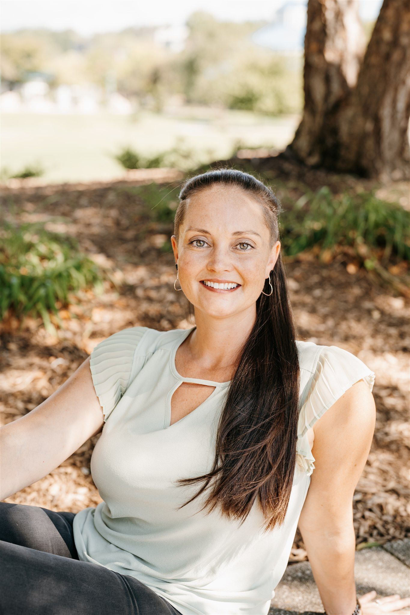 Woman with long dark ponytail smiles, seated outdoors in a park.