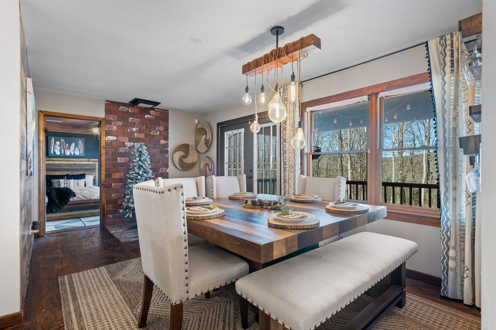 Dining room with wooden table, bench, chairs, and large window overlooking trees.