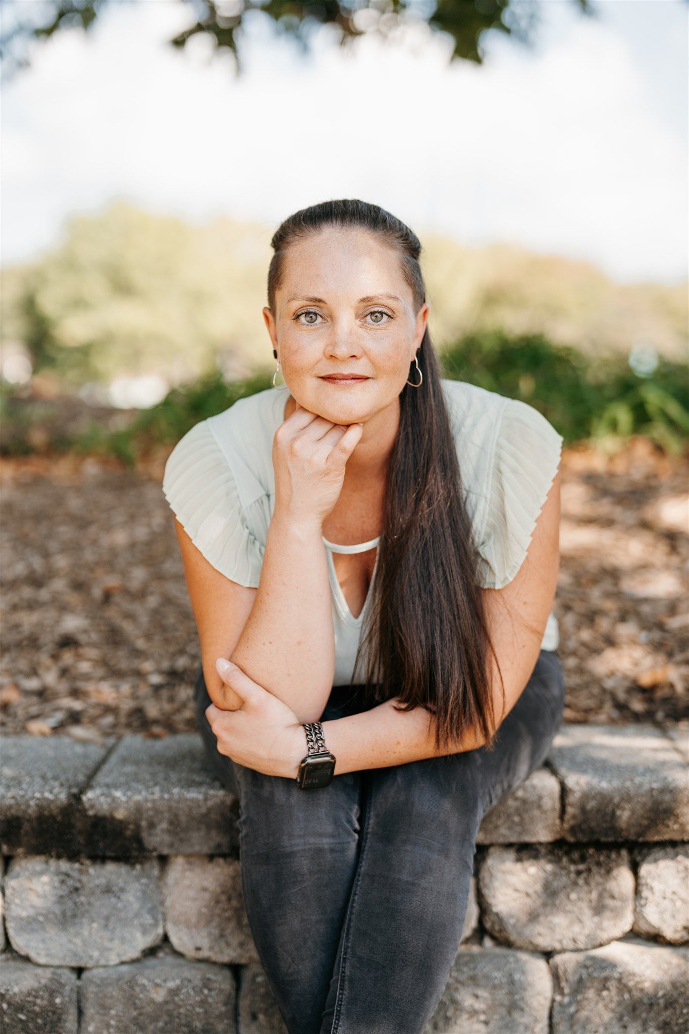 Woman seated outdoors, hand on chin, looking towards camera. Light green shirt, dark jeans, watch, long brown hair.