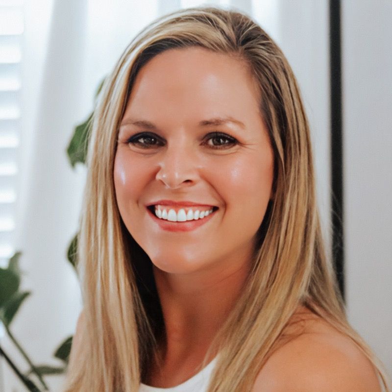 Woman with long blonde hair smiles, wearing a black jacket and gold necklace against a gray background.