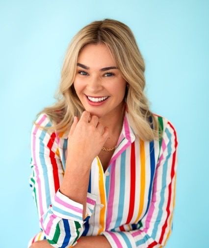 Woman with long blonde hair smiles, wearing a black jacket and gold necklace against a gray background.