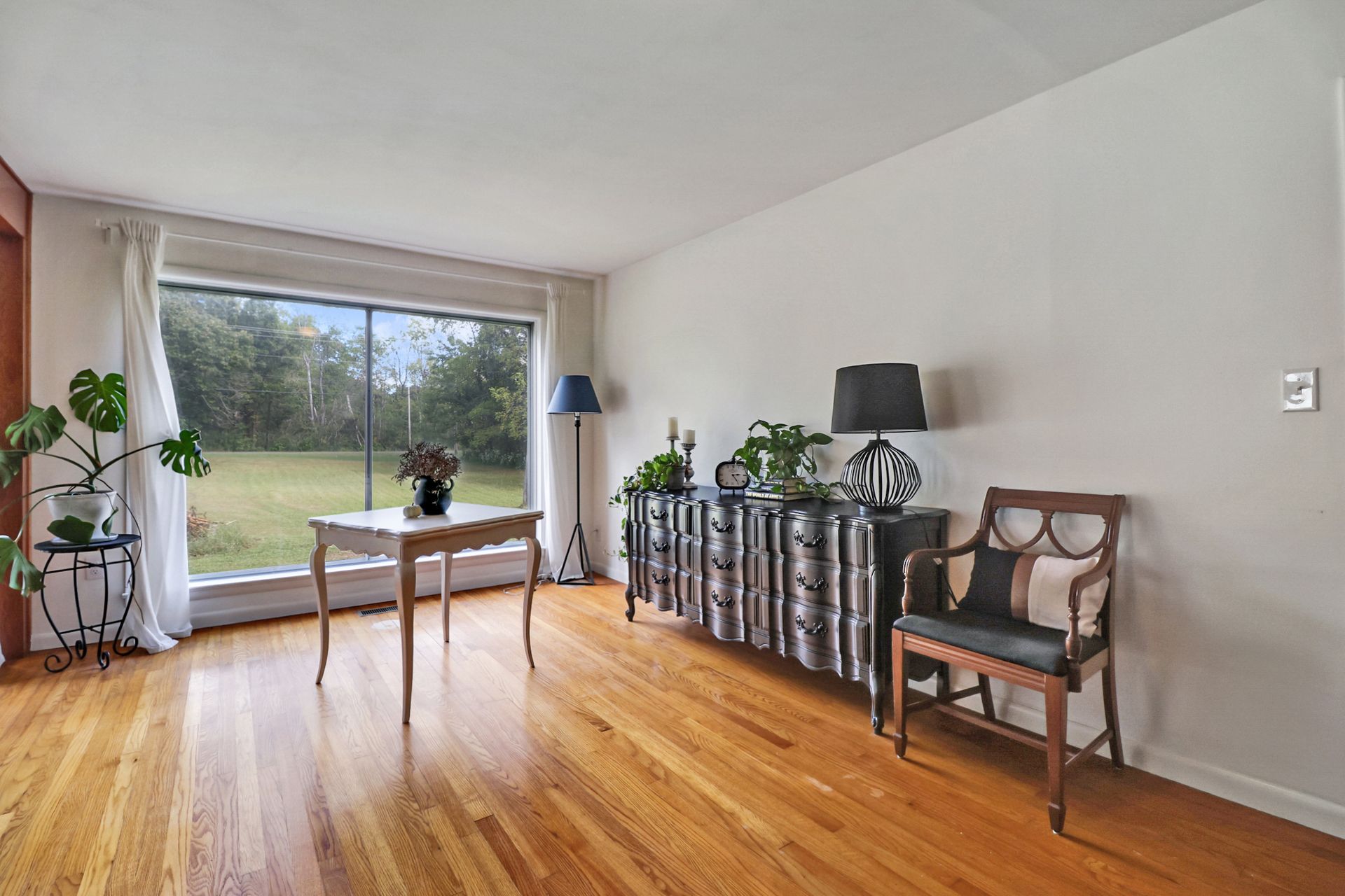 Living room with hardwood floors, a large window overlooking a yard, and a decorative cabinet.