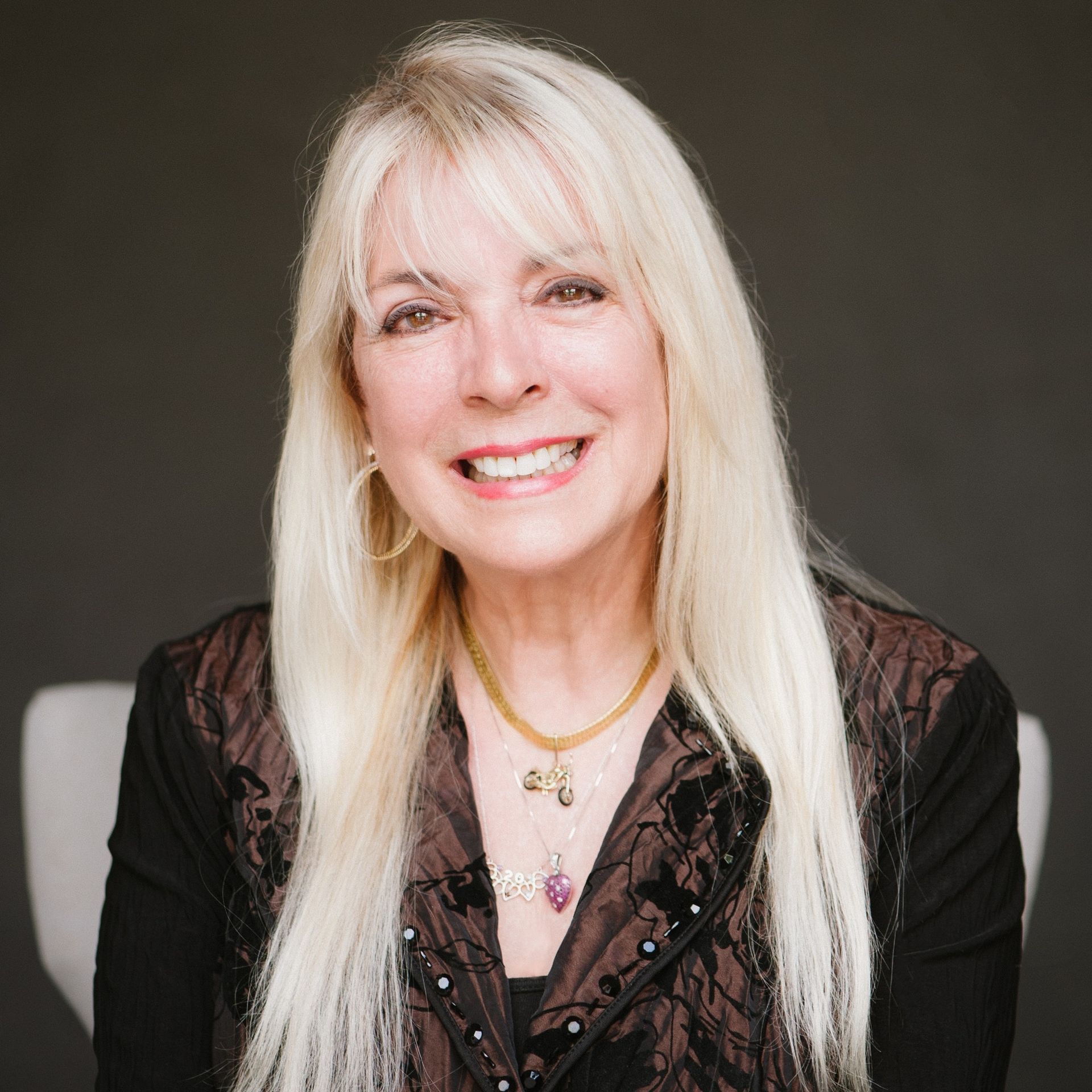 Woman with long blonde hair smiles, wearing a black jacket and gold necklace against a gray background.