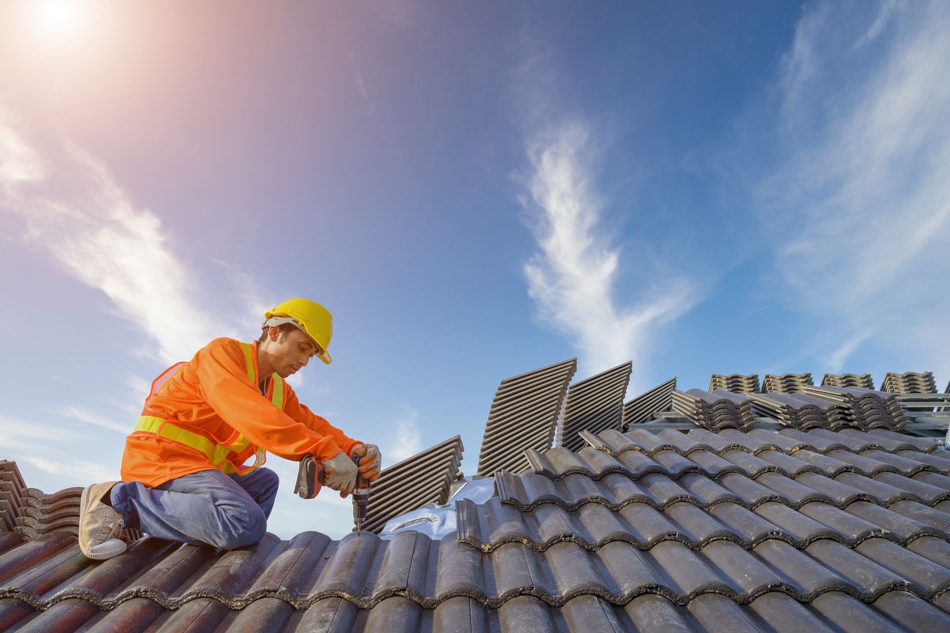 Roofer wearing safety gear, working on a tile roof against a blue, cloudy sky.