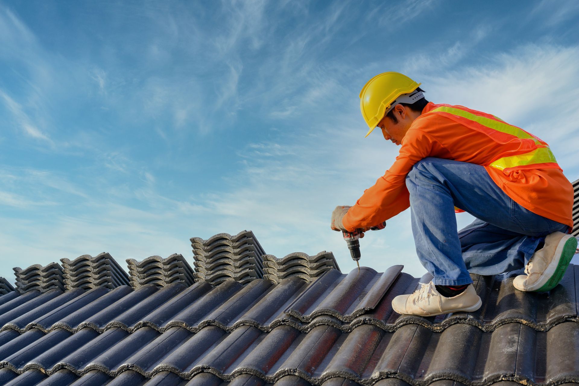Roofer in safety gear, installing dark ceramic tiles on a roof against a blue sky.