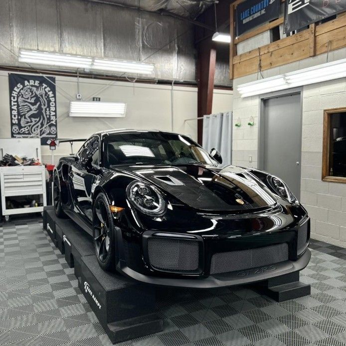 A black Porsche 911 GT2 RS sits on raised service ramps inside a professional auto shop with gray tiled flooring.