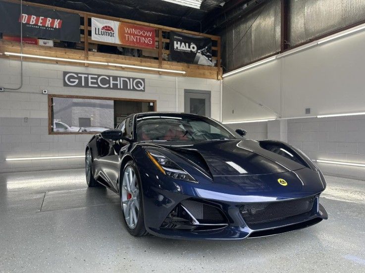 A dark blue Lotus Emira sports car parked inside a bright, professional detailing studio with Gtechniq signage.