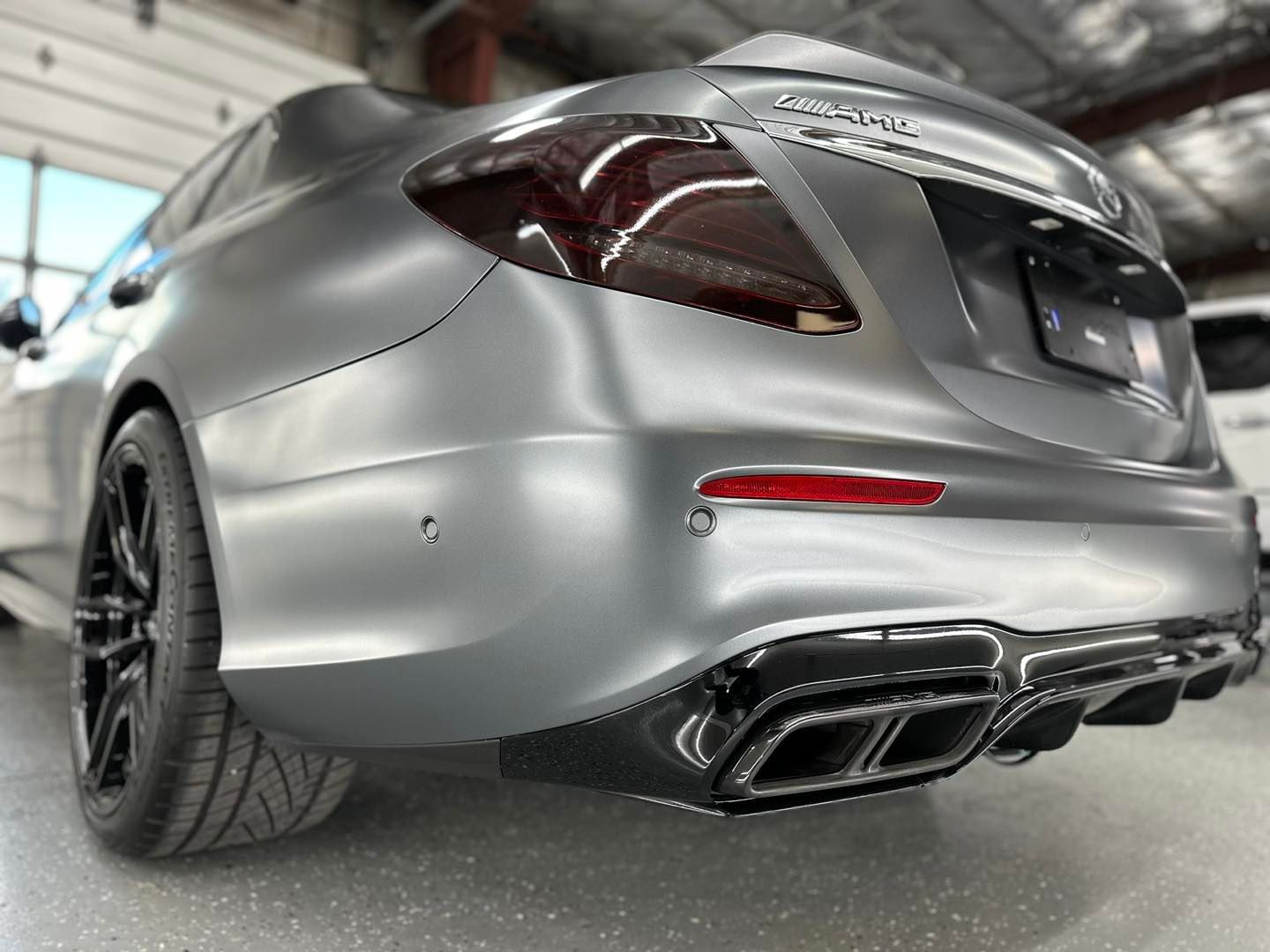 Rear view of a matte grey Mercedes-Benz AMG sedan parked in a garage, featuring black wheels and a carbon fiber diffuser.