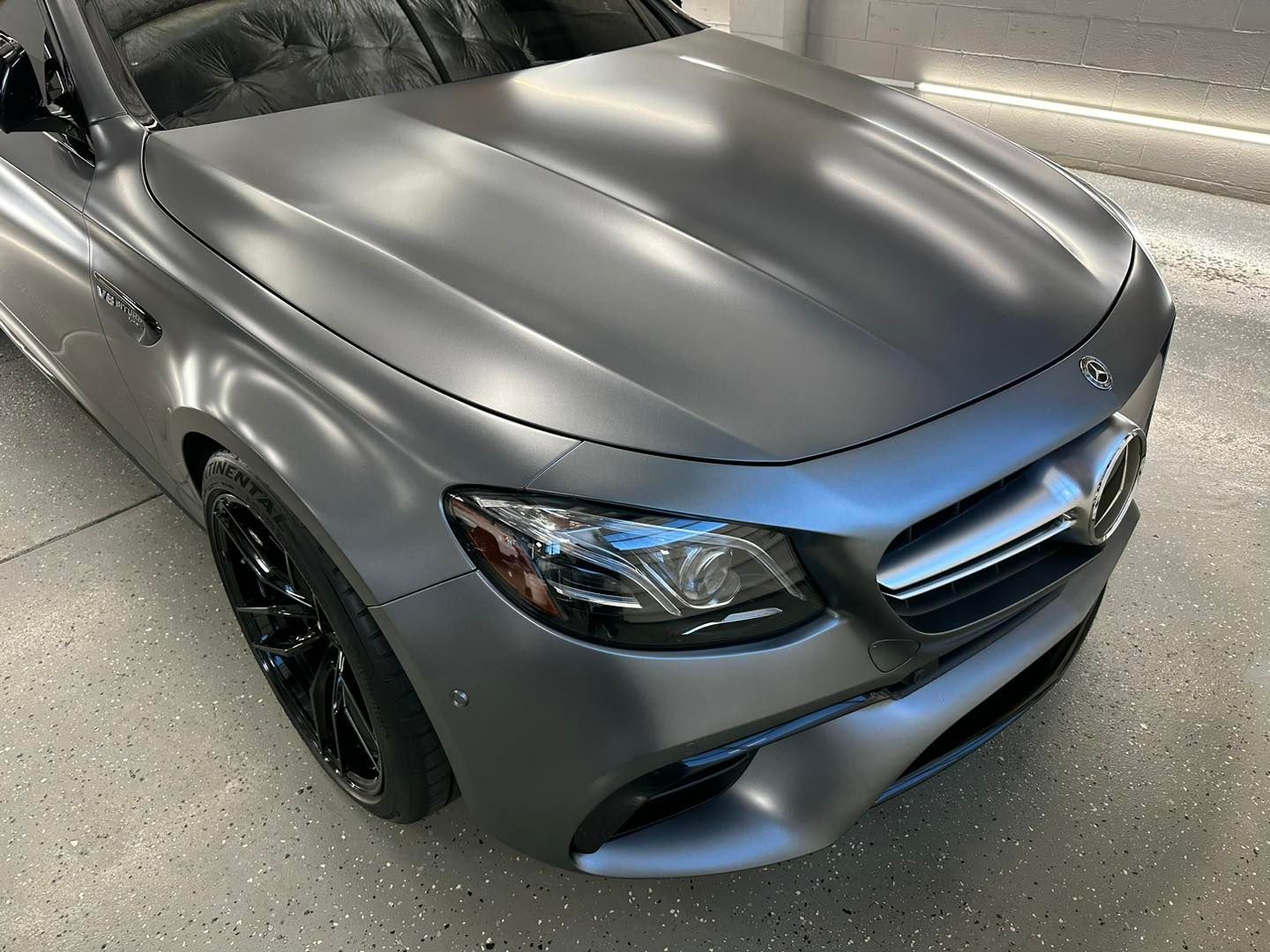 A matte gray Mercedes-Benz E-Class sedan parked on a speckled concrete floor, viewed from a high-angle front three-quarter.