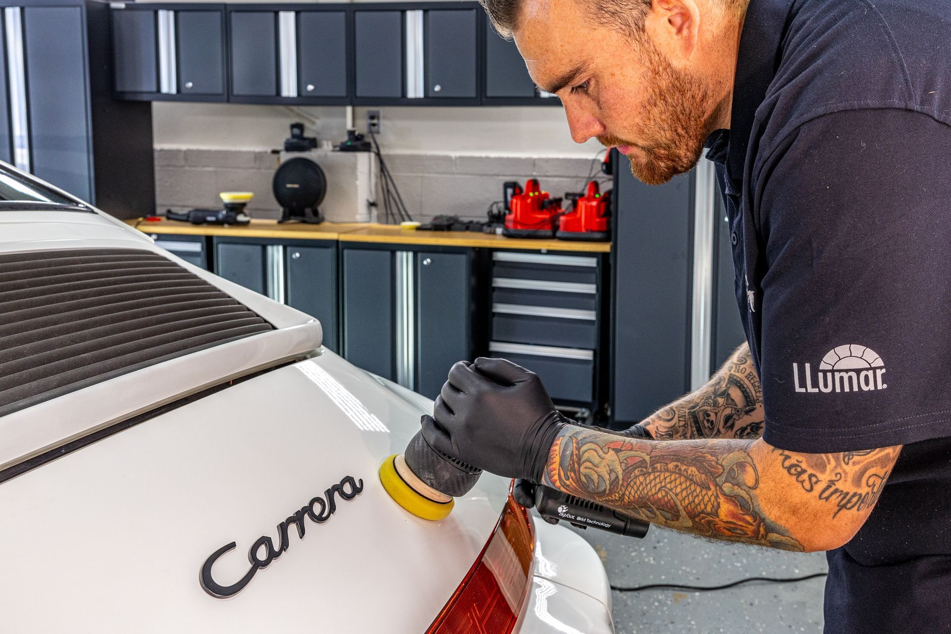 A man polishes a white Porsche Carrera in a garage, wearing gloves, with tools visible.