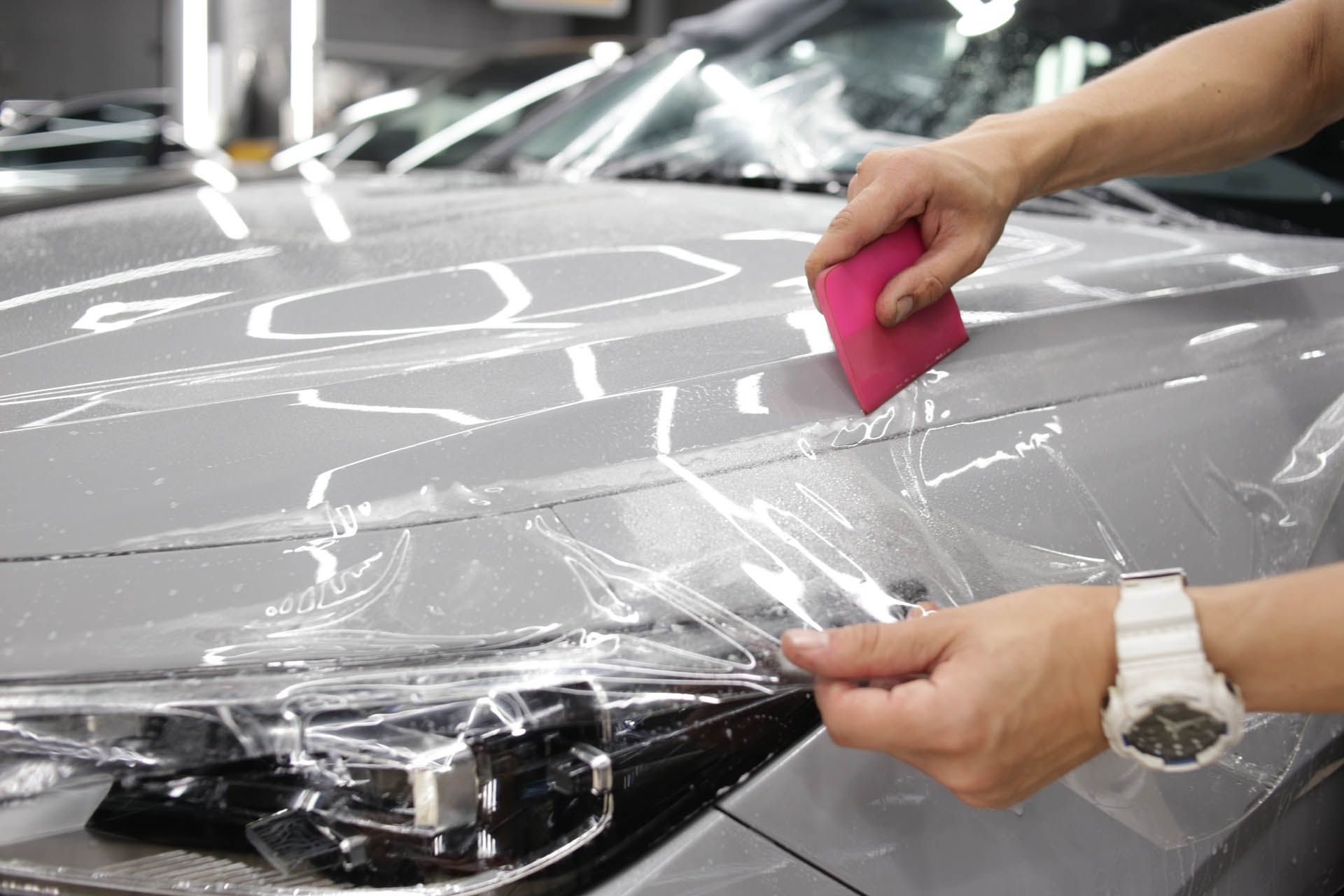 Person applying clear protective film to a gray car's hood, using a pink squeegee.