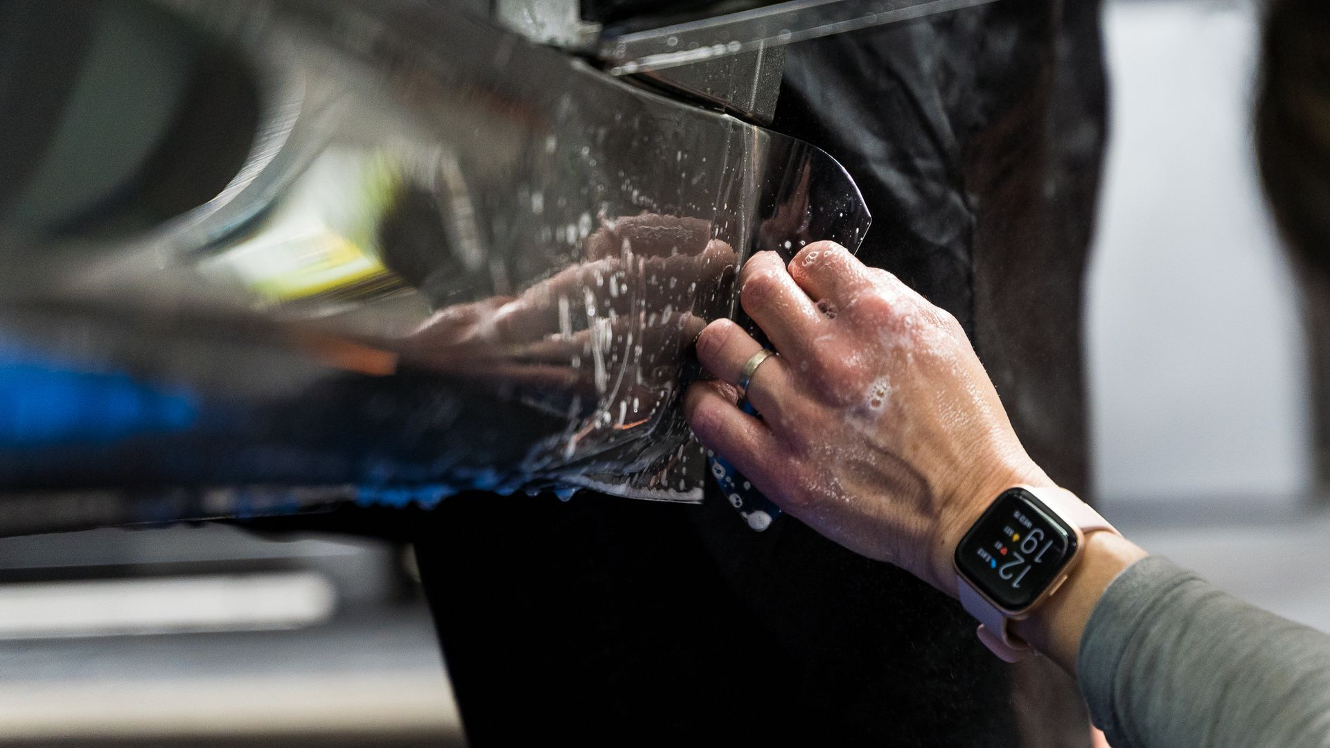 Person applying film to a car, wearing a smartwatch, in a garage.