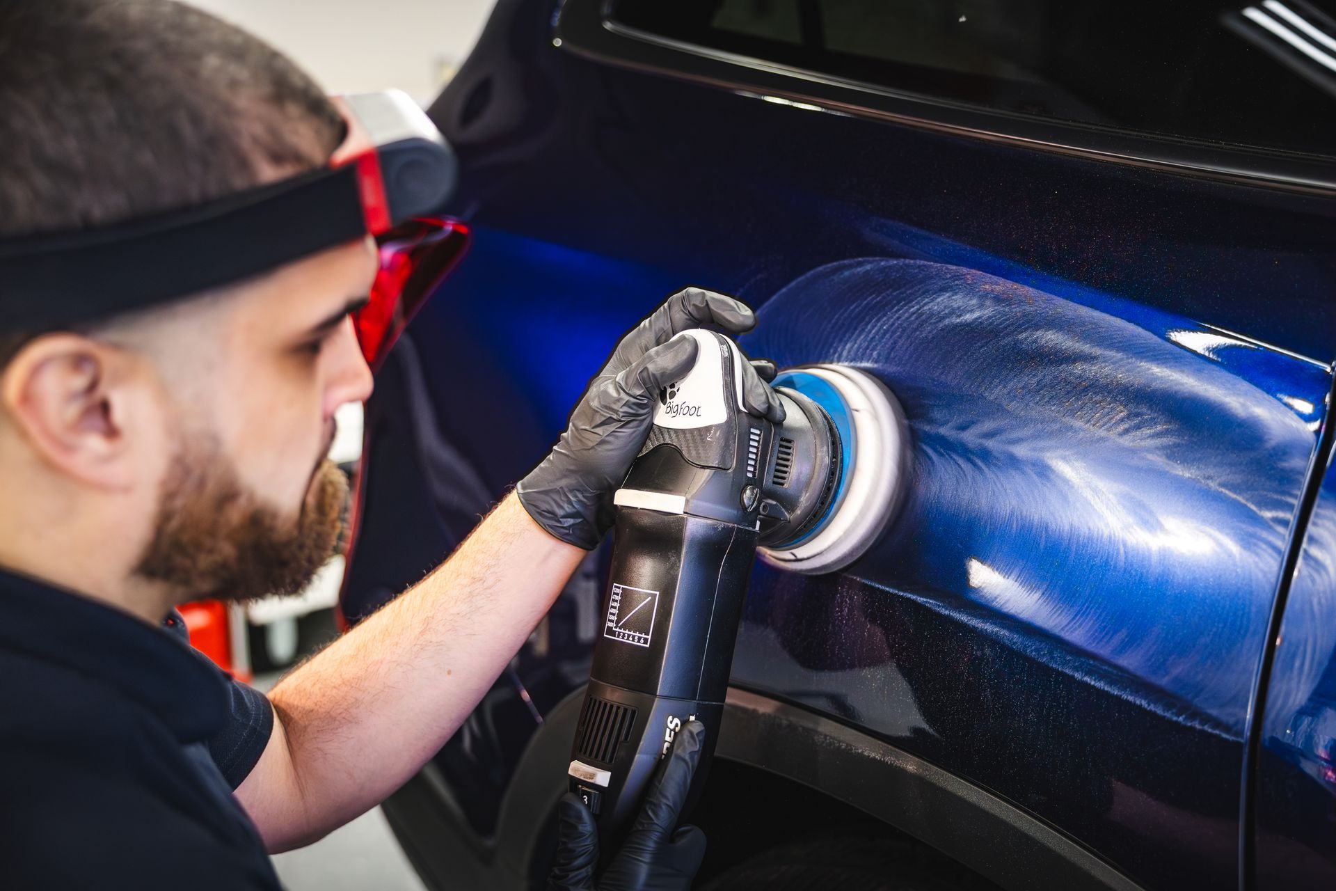 Man polishing a blue car with a machine, wearing gloves and a headlamp.