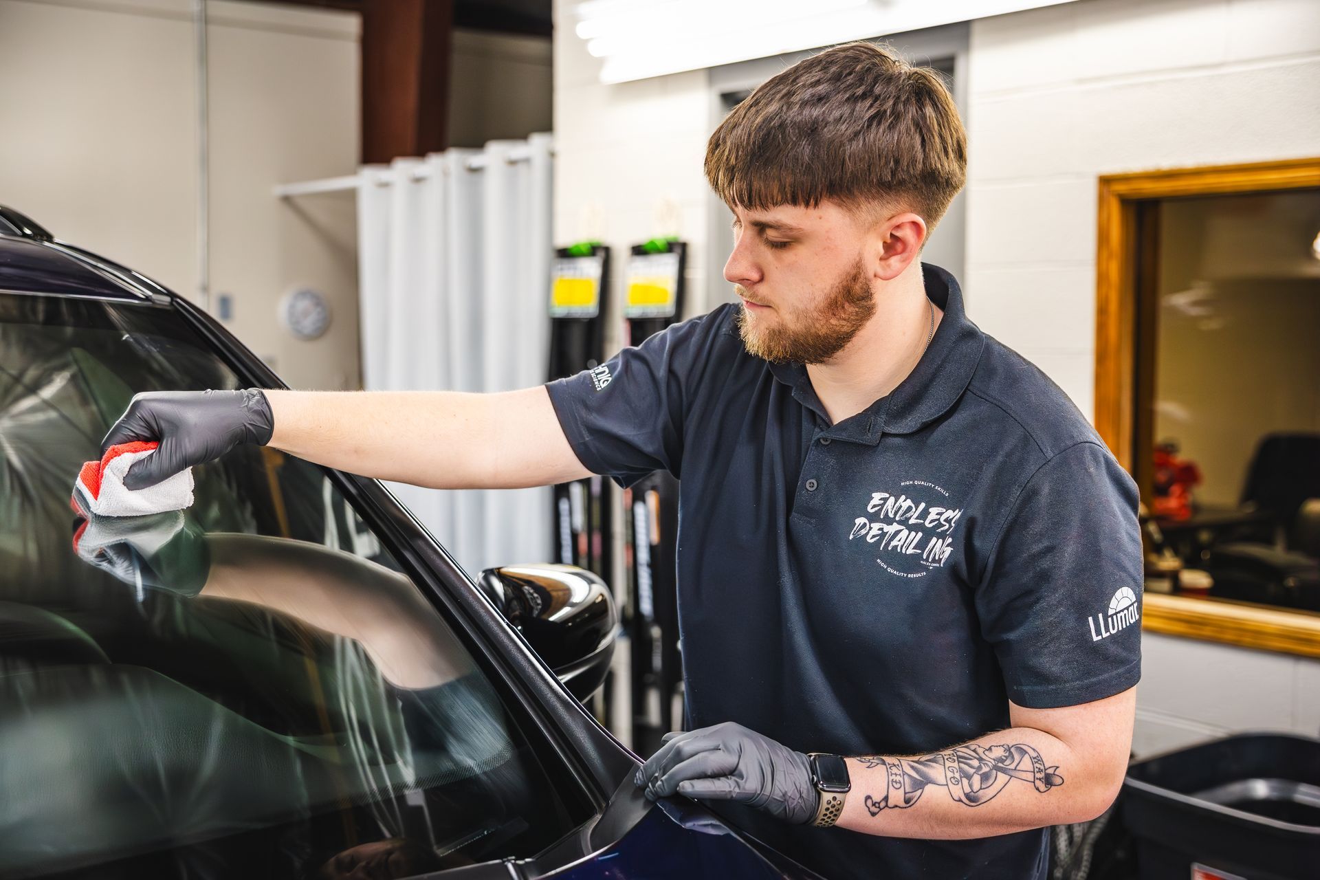 Man in black gloves, wiping windshield with a red applicator. Dark-blue shirt, indoor setting.