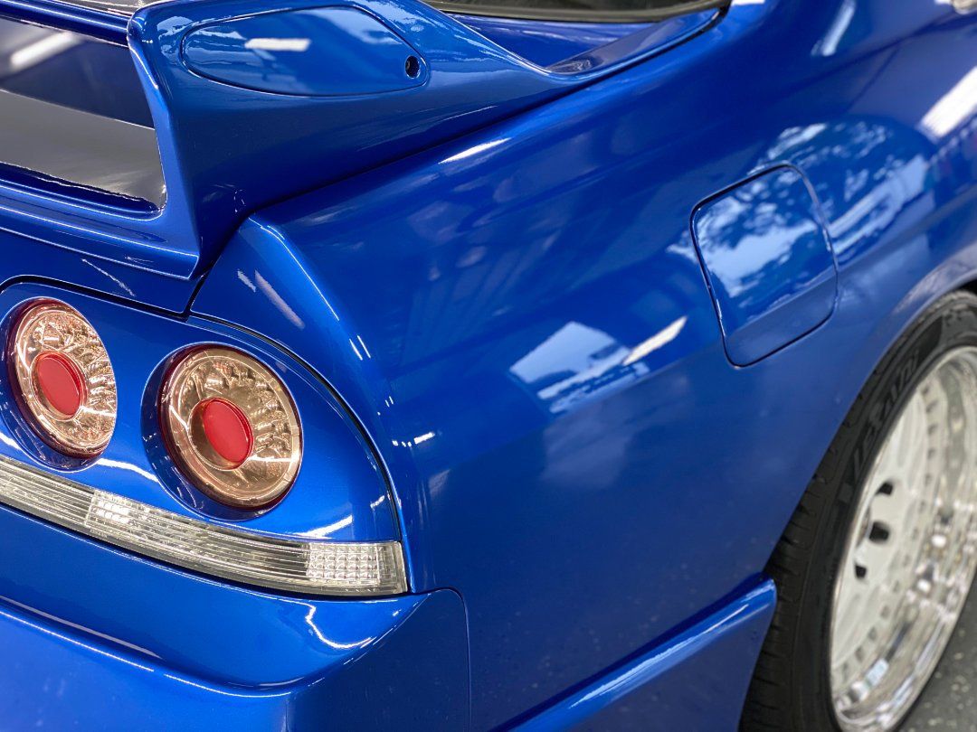 Close-up of a blue sports car’s rear quarter panel with twin round taillights and a chrome wheel