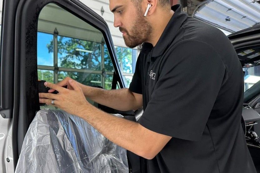 A man applies tint to a car window in a garage. He wears a black shirt and has an earpiece.