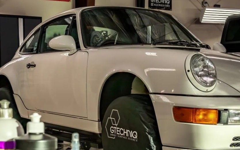 White Porsche 911 coupe in a shop, with headlight and tire cover visible.