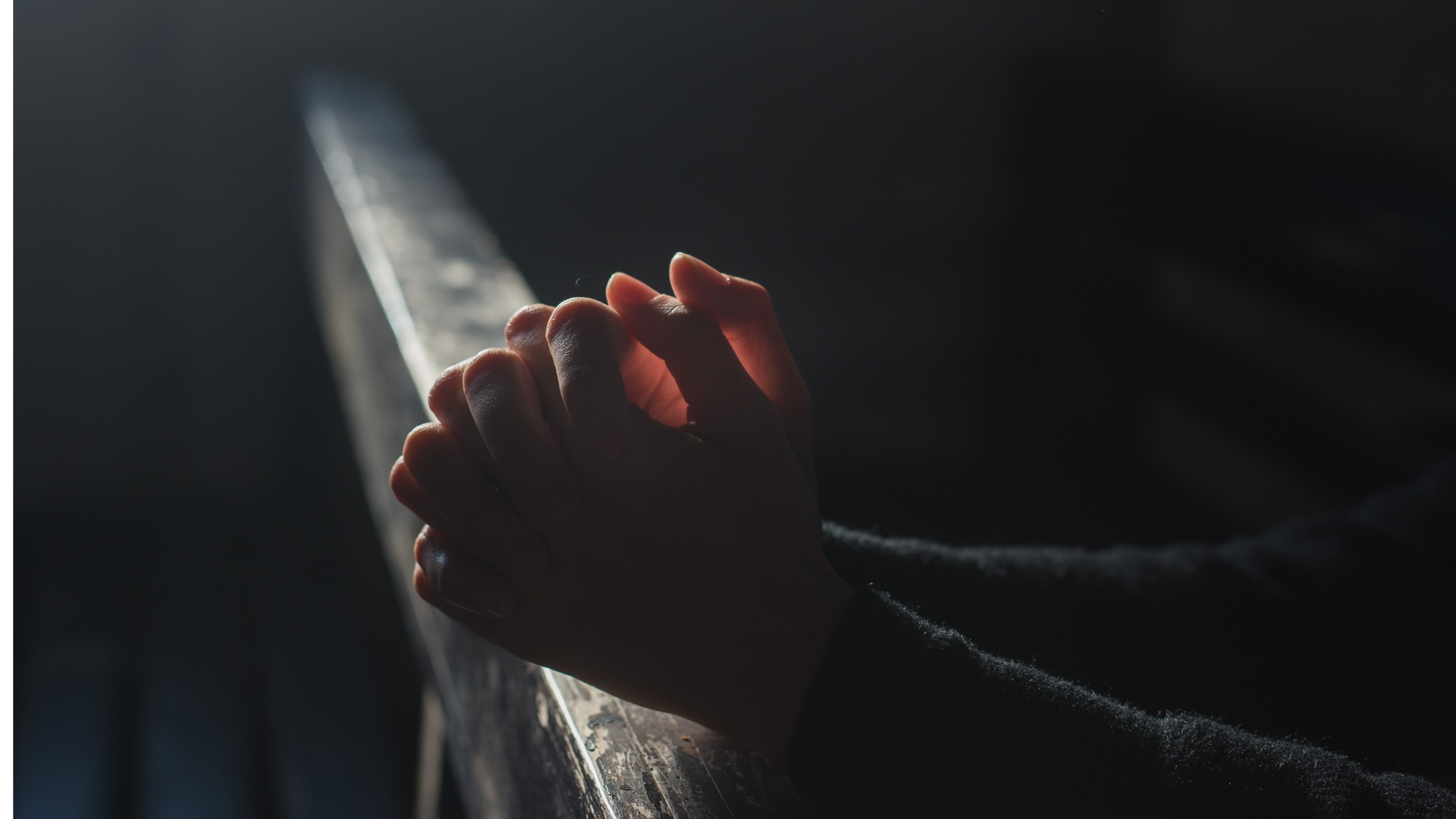 A person is praying with their hands folded in a dark room.