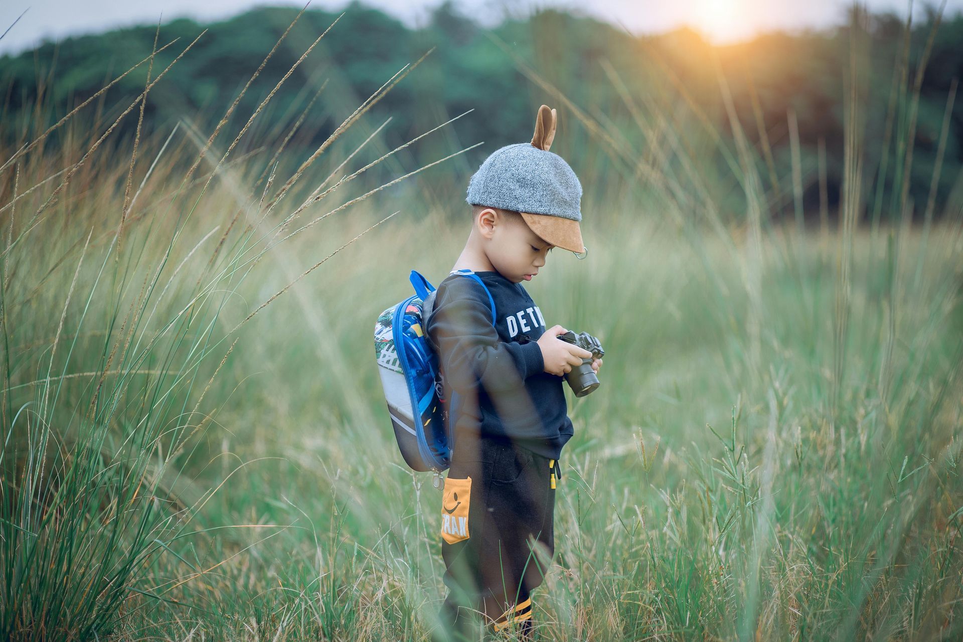 A young boy with a backpack is standing in a field holding a camera.