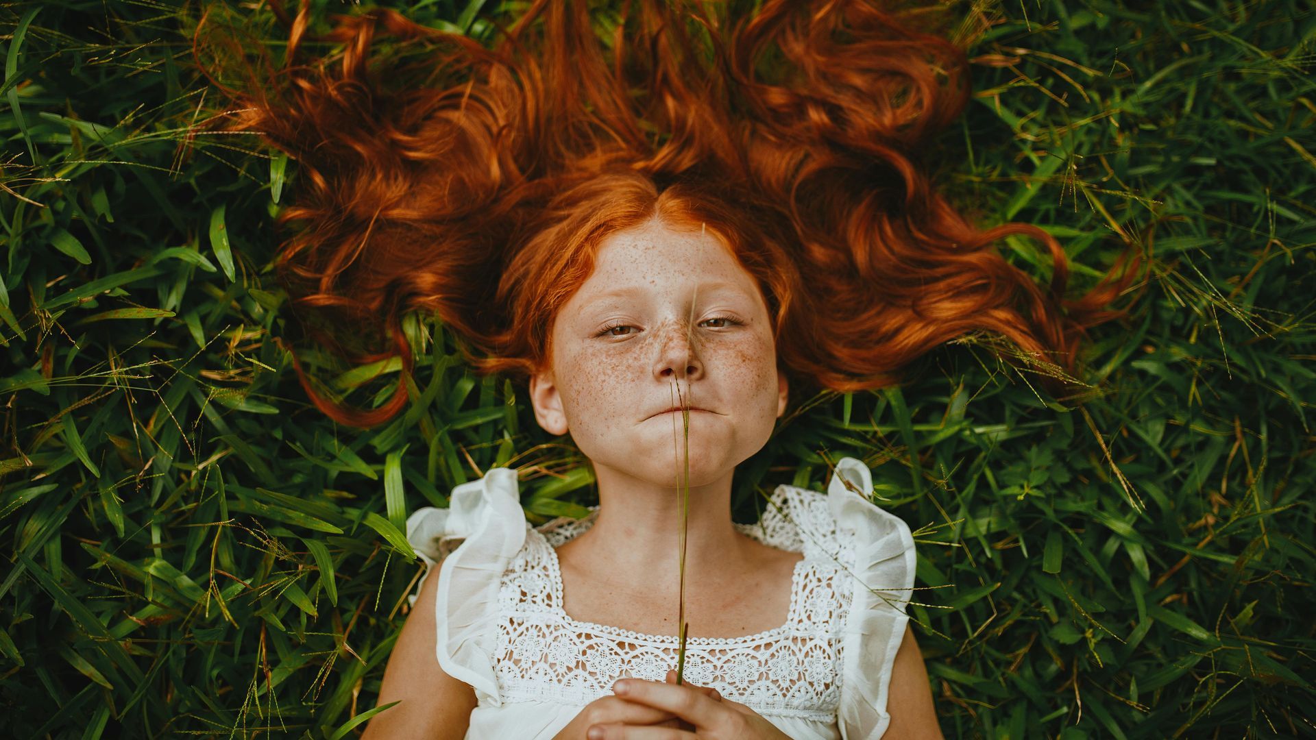 A little girl with red hair is laying in the grass with a flower in her mouth.