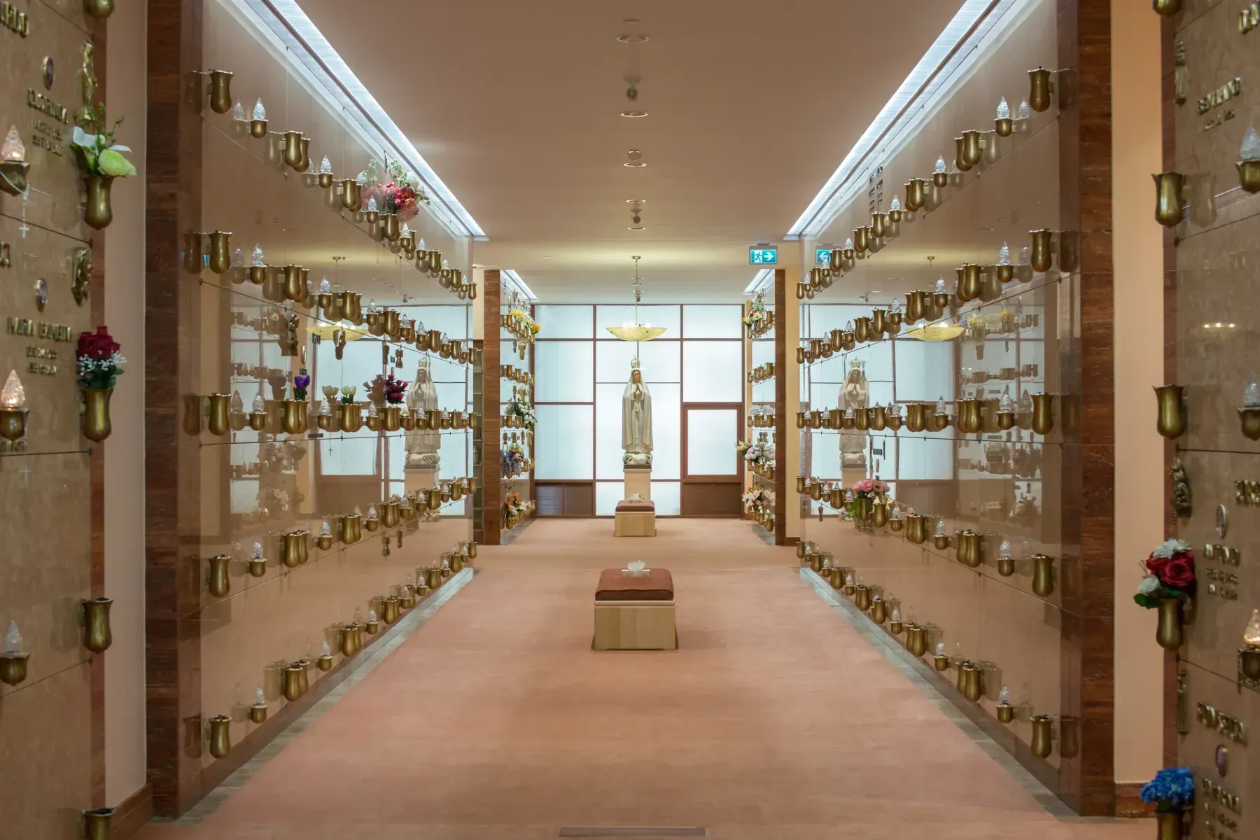 Wide corridor in a columbarium, with rows of niche plaques and flowers lining both walls.
