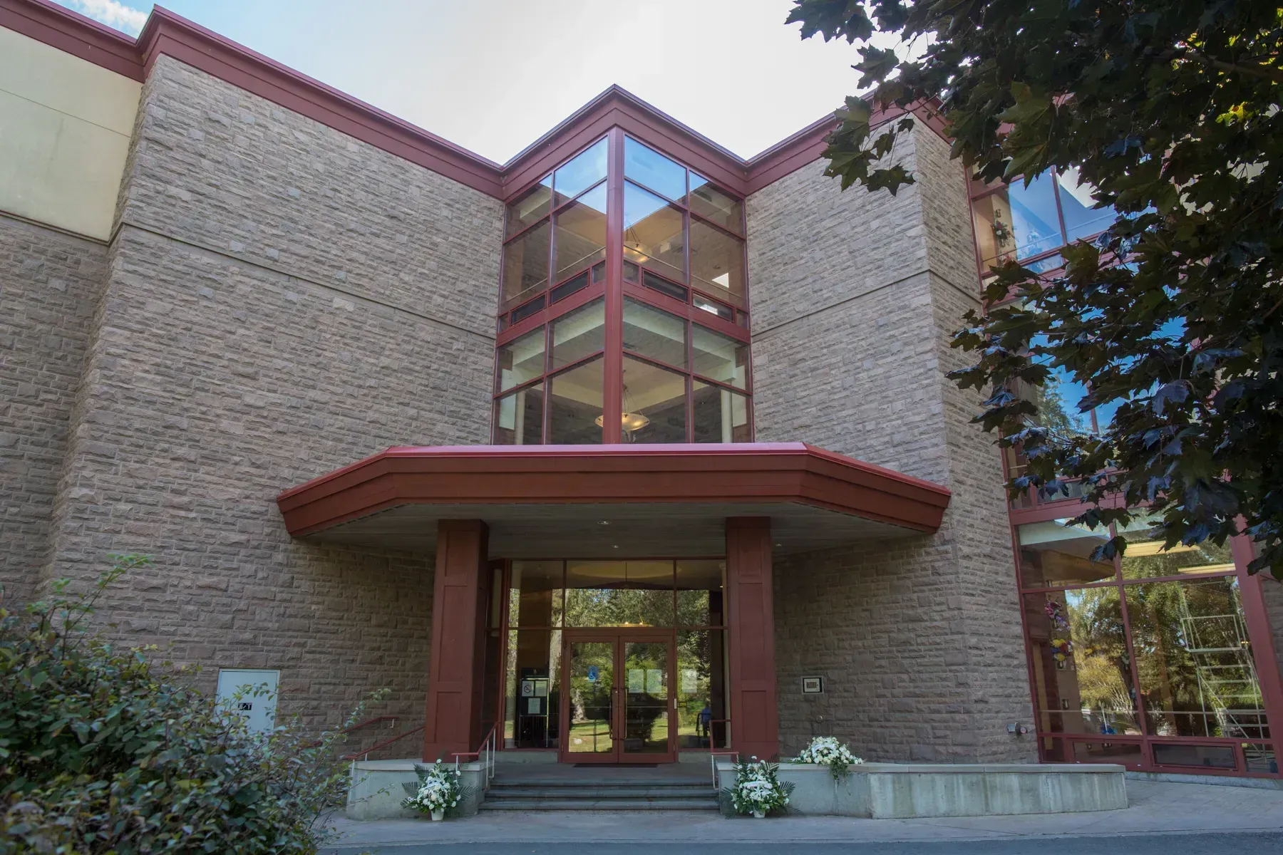 Modern brick building entrance with glass atrium and red trim, surrounded by trees and flowers