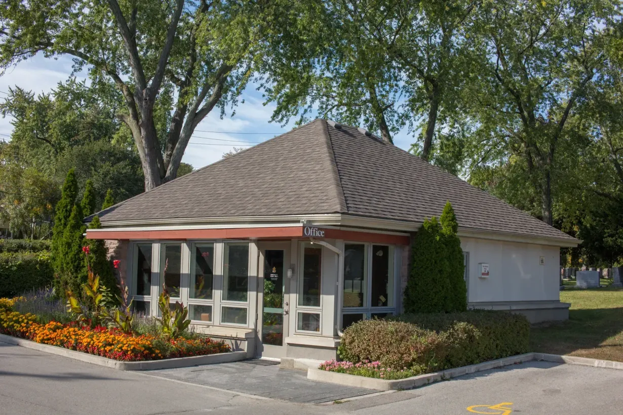 Small building with glass entrance, red awning, and flower beds surrounded by trees.
