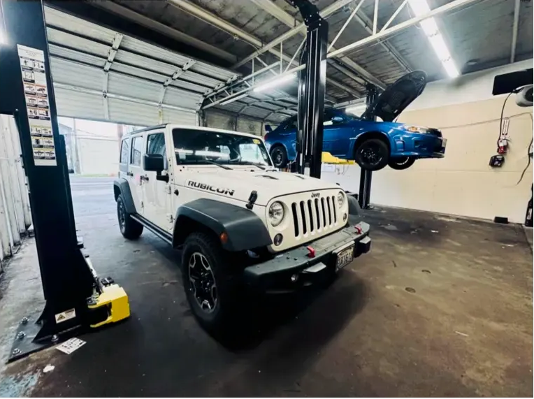 White Jeep Wrangler and blue car on a lift in a garage. | Supreme Auto Repair Shop