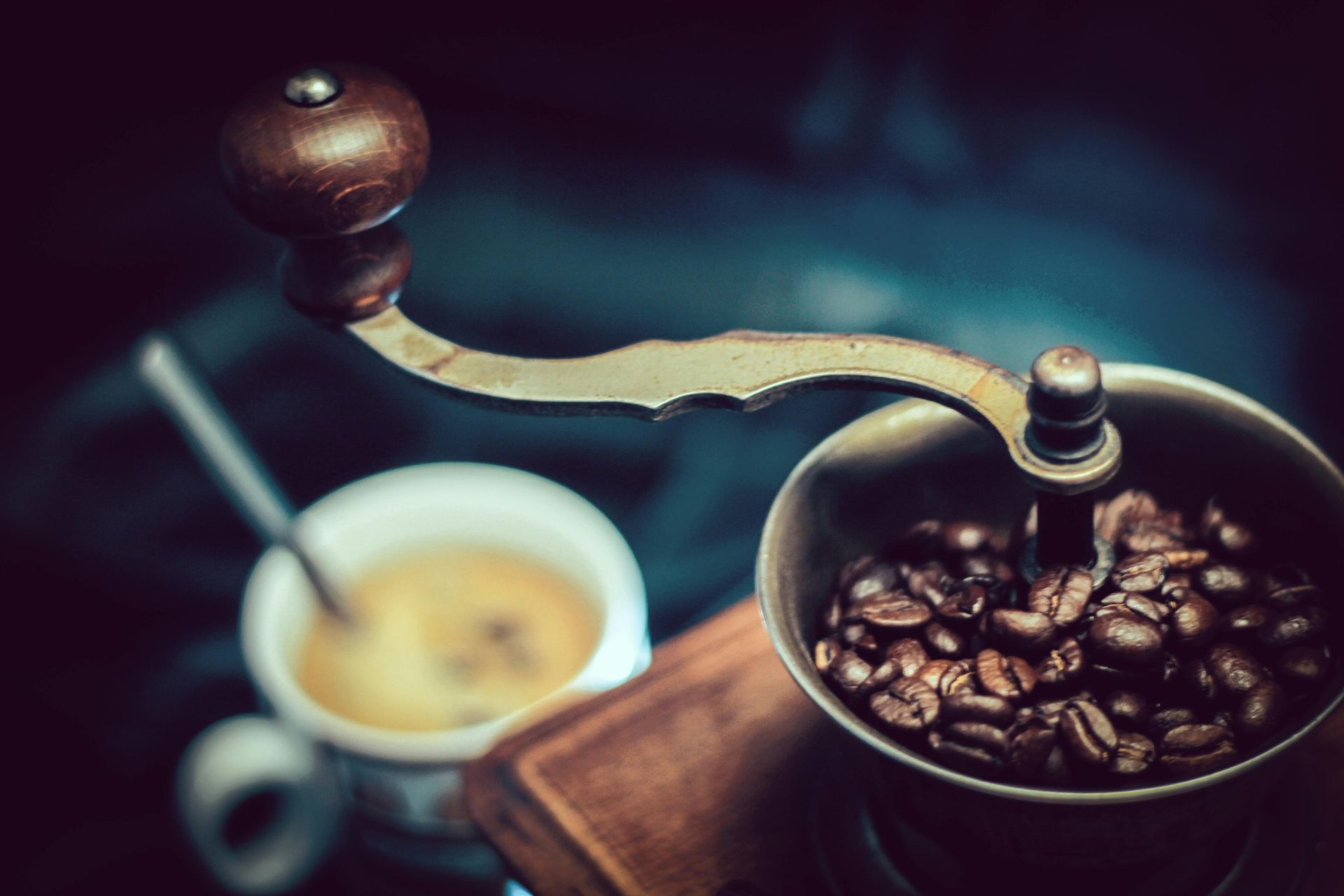 Coffee grinder with beans, mug of coffee, and spoon on dark surface.