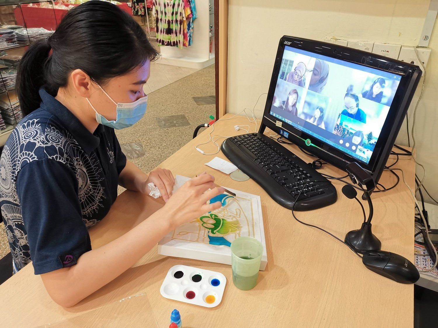 Woman in mask paints, participating in a virtual art class. Paints, water, and a computer with other participants on a table.