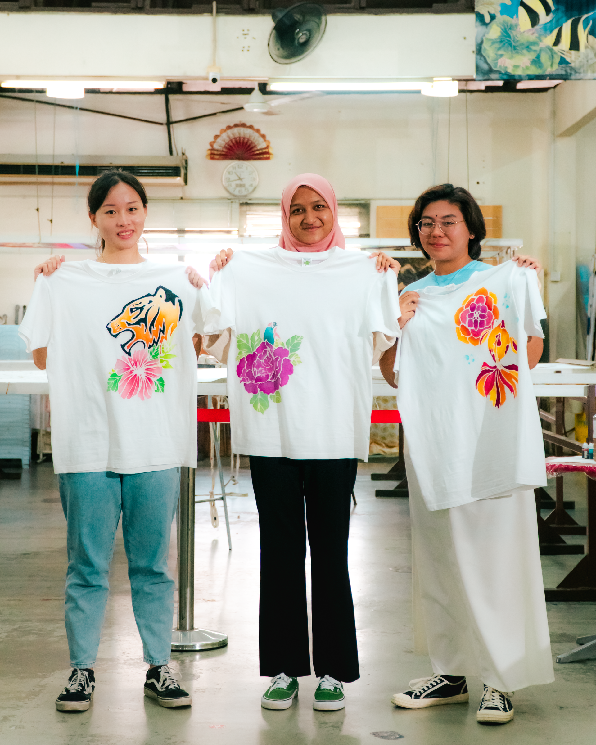 Three women holding up white t-shirts with colorful floral designs, smiling in a workshop.