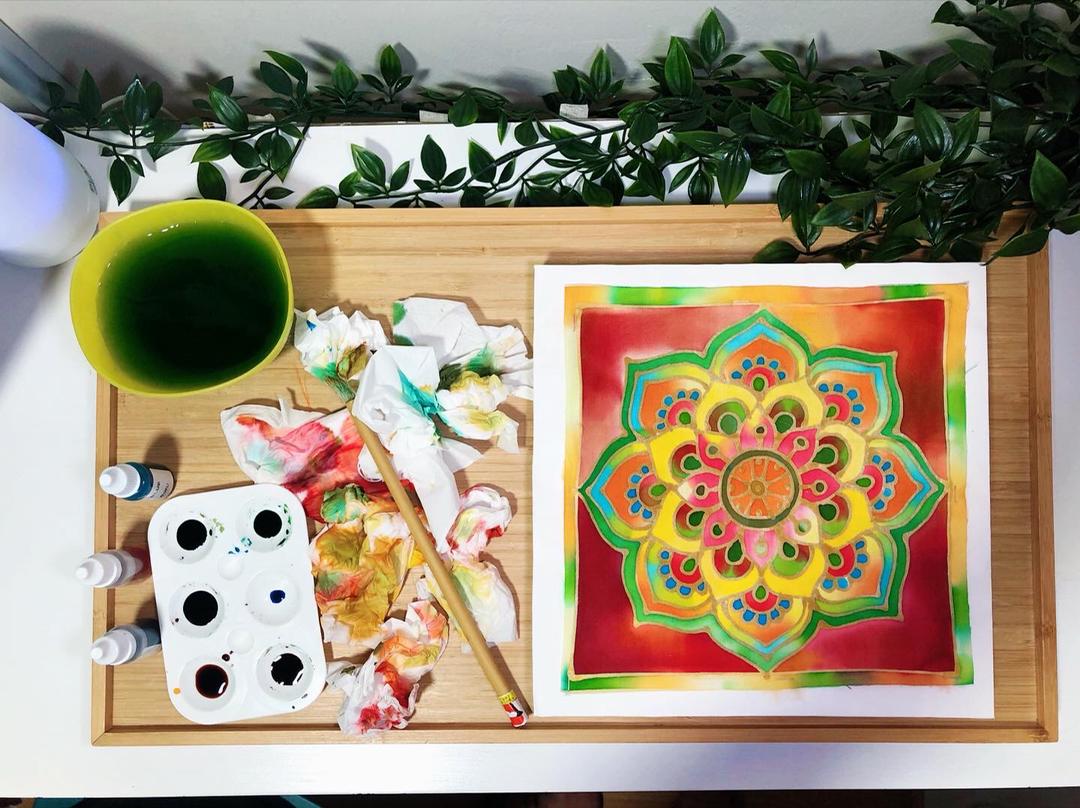 Wooden tray with vibrant mandala art, paint, brush, and green water; setting with green foliage.