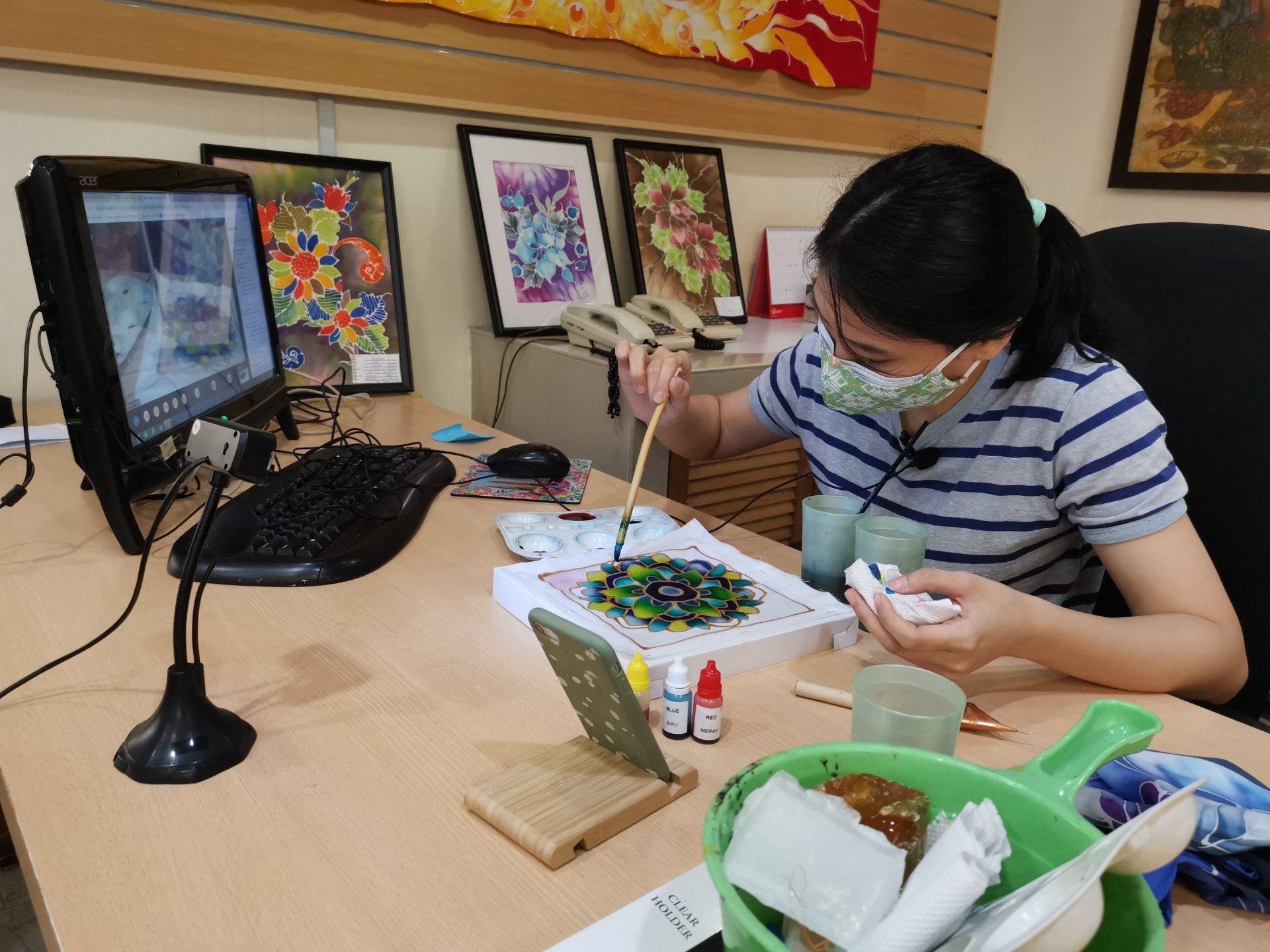 Woman wearing a mask painting colorful artwork at a desk, computer on the left, framed art in the background.