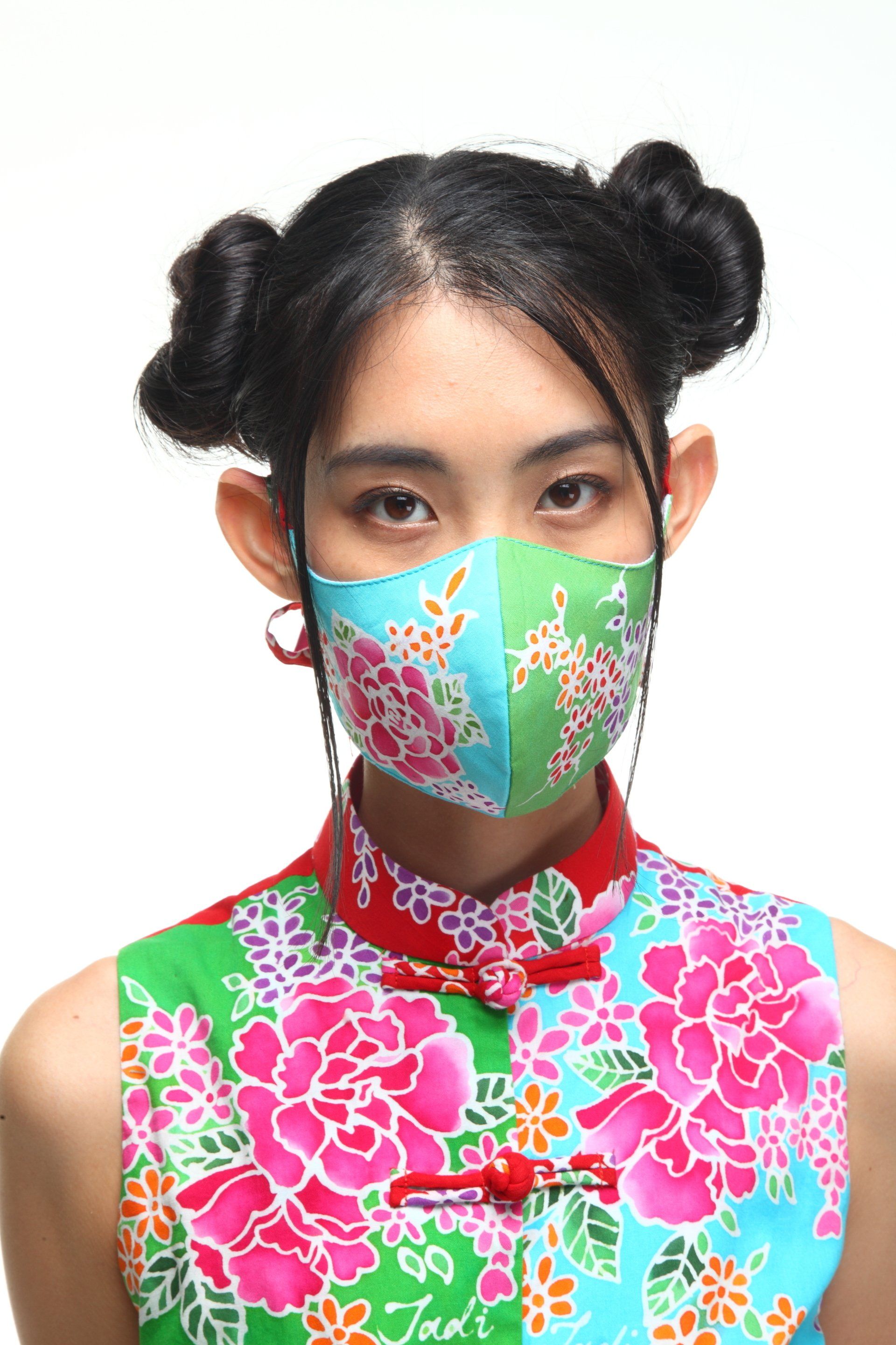 Woman in floral outfit and mask, hair in buns, against a white background.