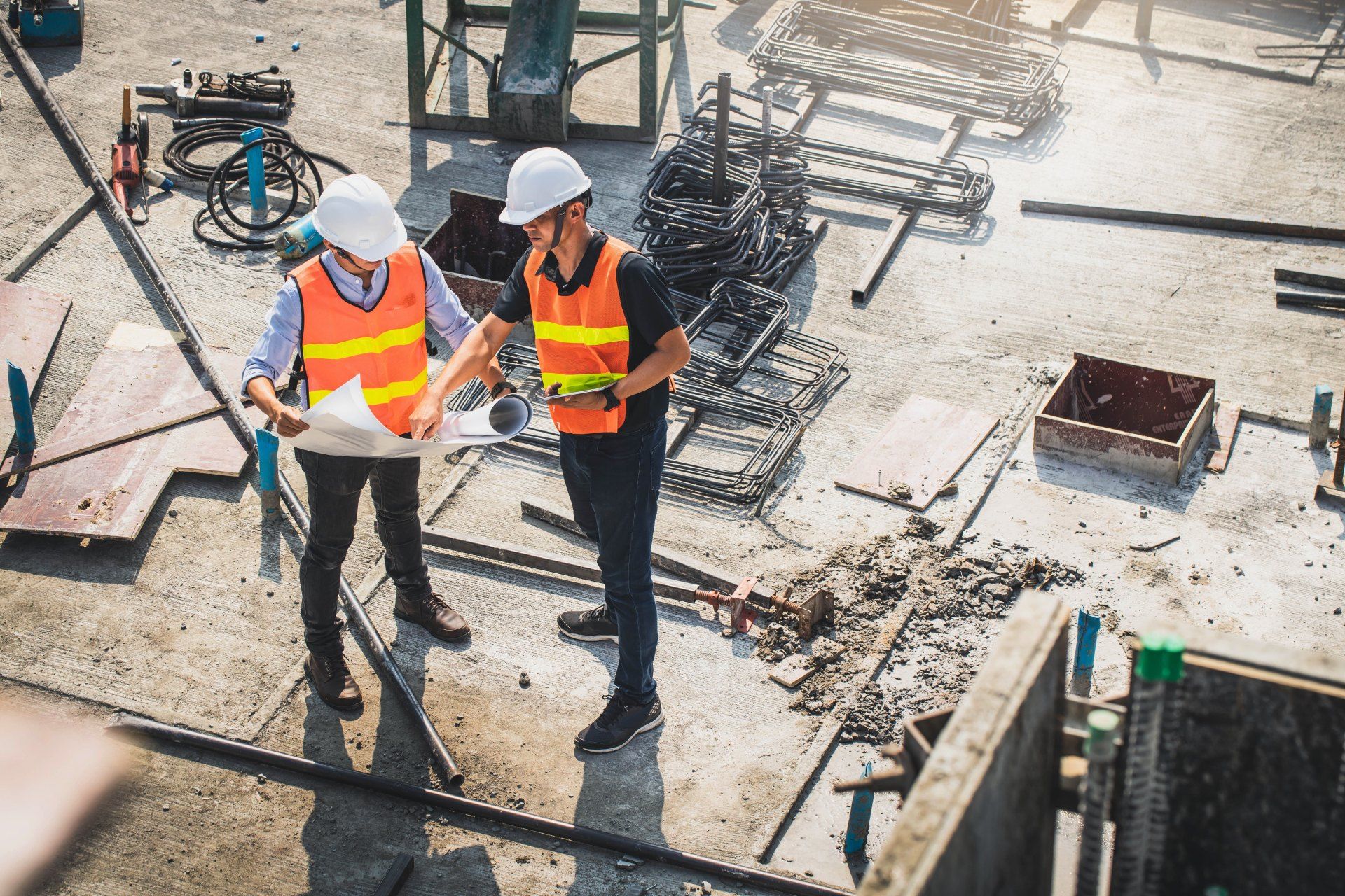 Two construction workers are standing on a construction site looking at a blueprint.