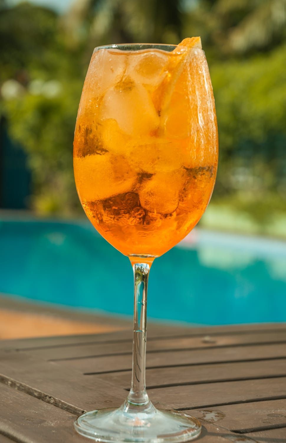 Um copo de suco de laranja com cubos de gelo está sobre uma mesa de madeira ao lado de uma piscina.