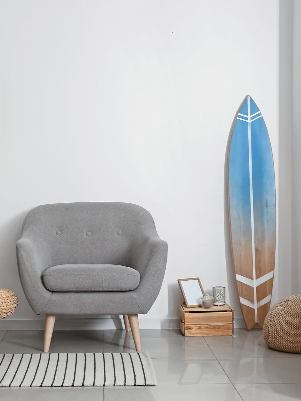 Gray armchair and a decorative surfboard against a white wall; rug and small wooden crate.