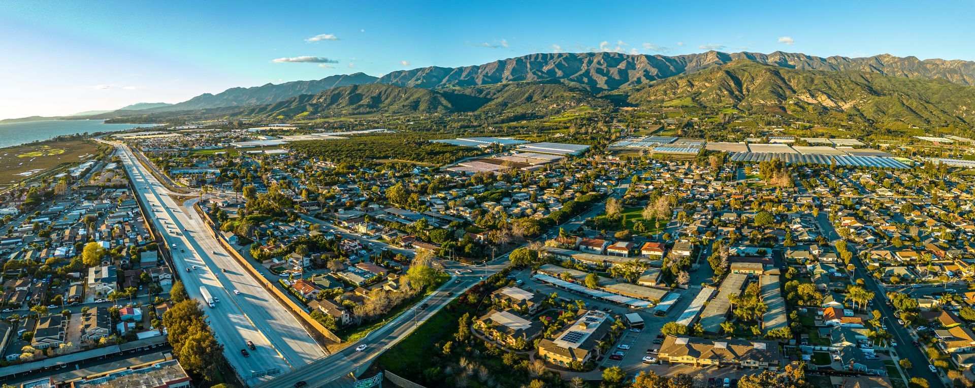 Aerial view of a city with a highway, mountains, and a body of water in the background, under a blue sky.