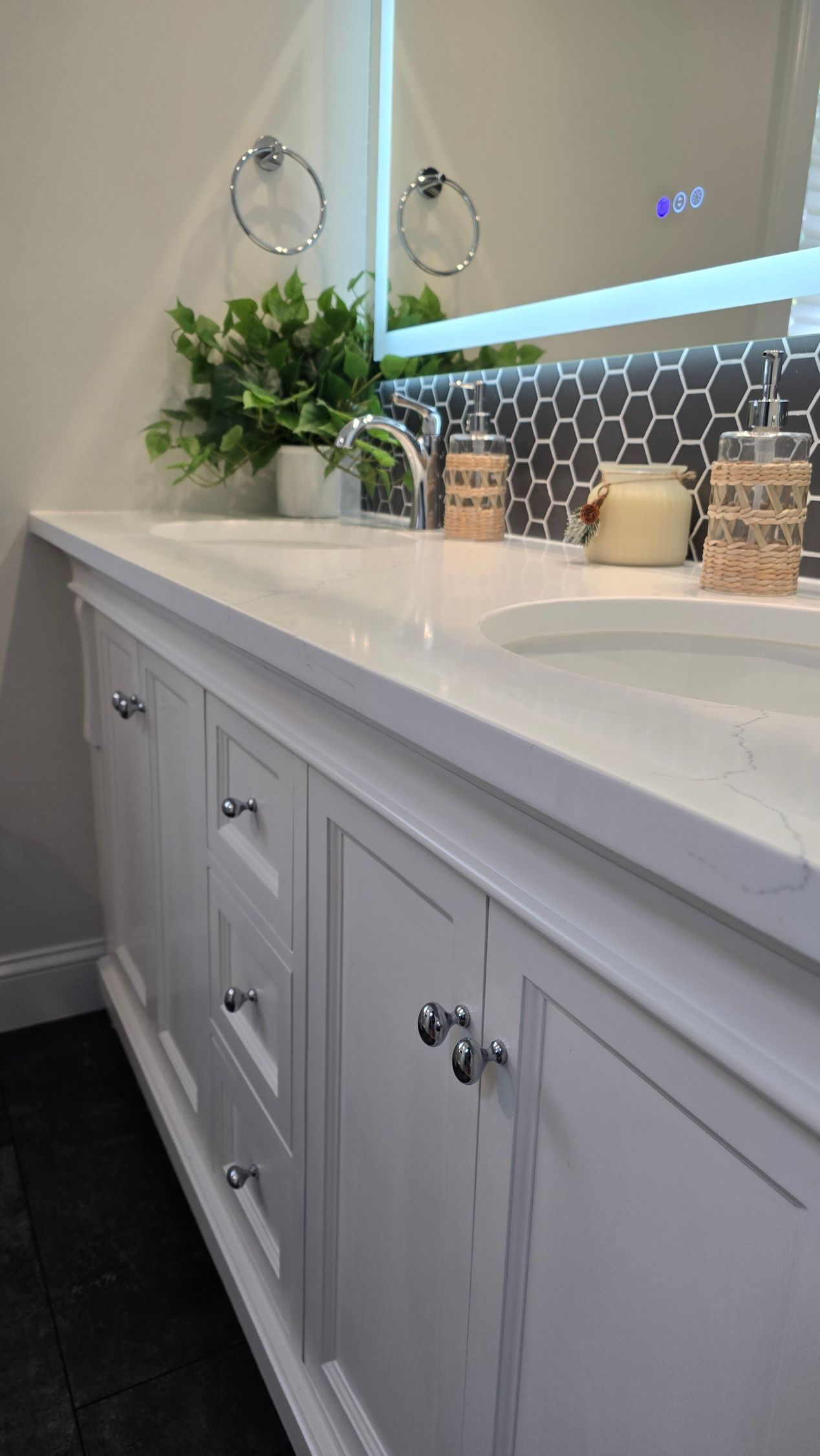 This image showcases a beautifully designed bathroom remodeled in West Hartford by First Choice Bathroom Remodeling with a bright and elegant aesthetic. The space features a modern white vanity with gold hardware, clean lines, and a minimalist open shelf for storage. A round mirror above the vanity is flanked by two wall-mounted sconces, providing soft, warm lighting.
The deep soaking tub is adorned with classic white subway tiles, creating a timeless and clean look. The gold fixtures, including a luxurious rainfall showerhead and handheld sprayer, add a touch of sophistication and warmth to the space. The hexagonal mosaic floor tiles introduce texture and contrast, complementing the overall design.
This bathroom blends modern luxury with classic elegance, making it feel both stylish and inviting. The gold accents stand out beautifully against the crisp white backdrop, giving the space a fresh, high-end appeal.