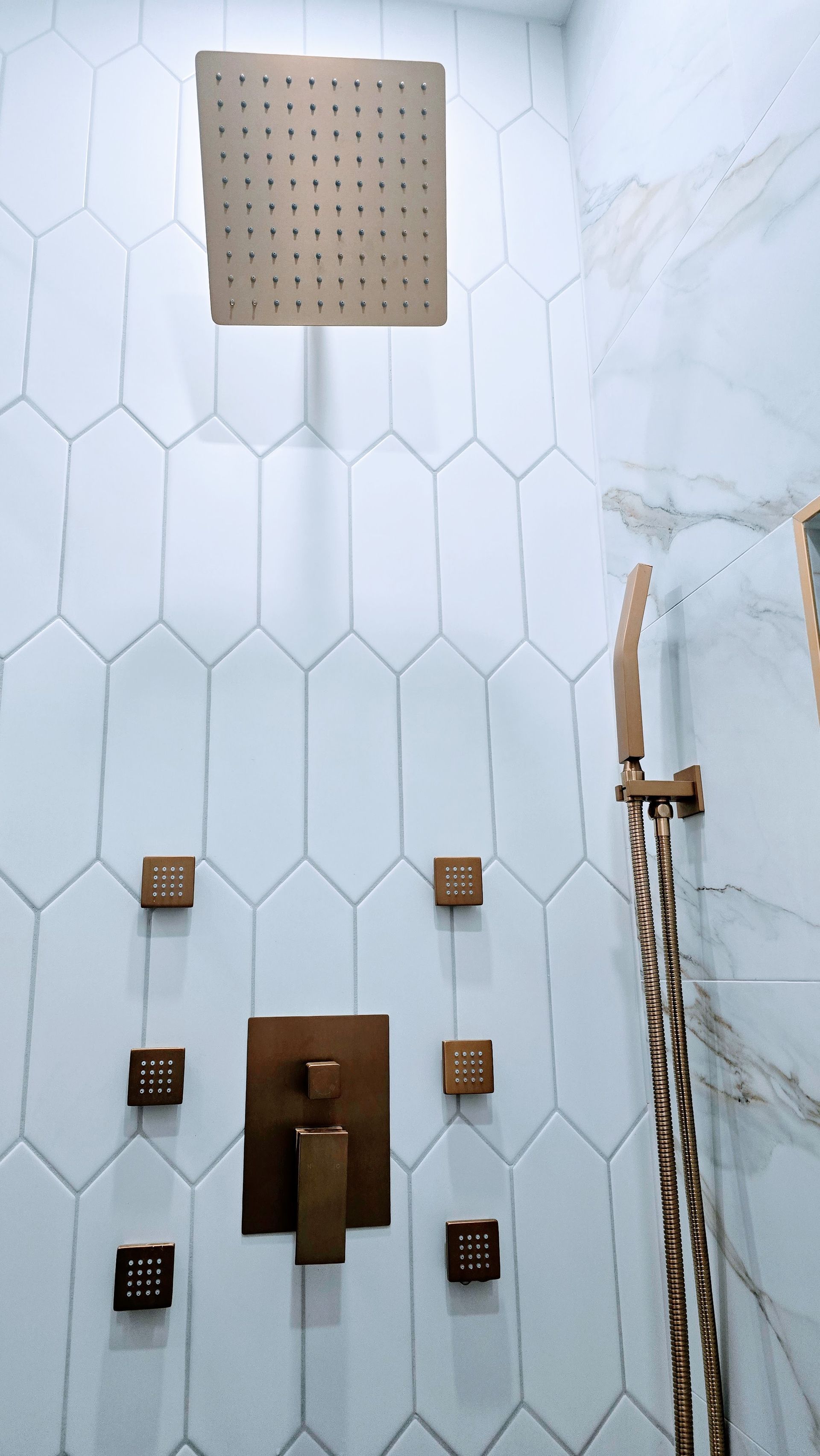 Shower interior with bronze fixtures, including a rain showerhead, hand shower, and water controls on a white tiled wall.