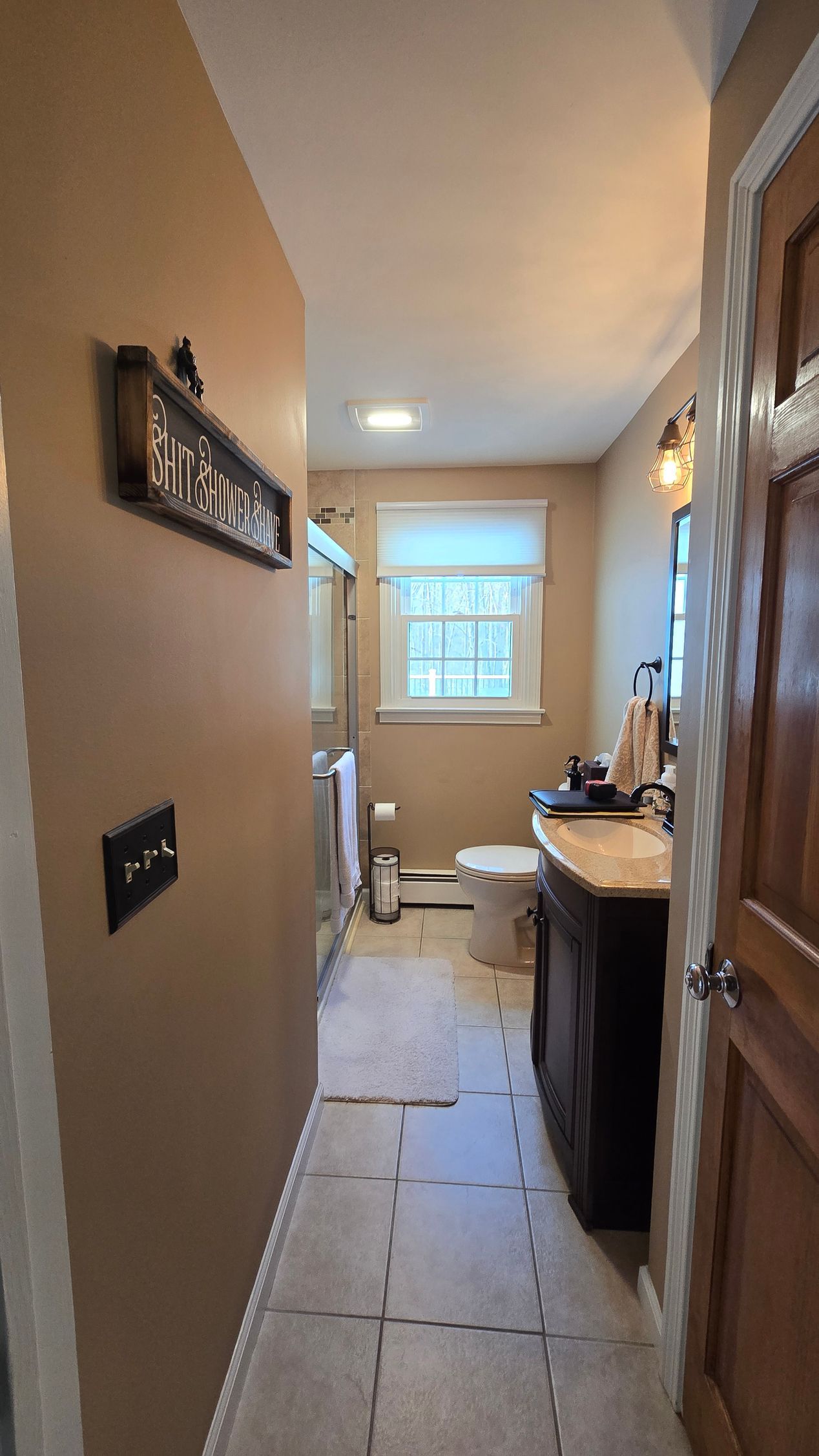 Narrow bathroom with dark vanity, toilet, and window, tan walls, and light tiled floor.