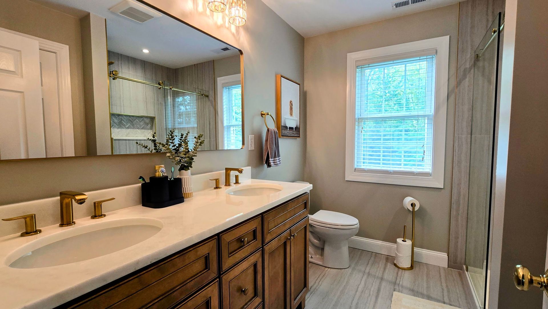 A bathroom with a double vanity in Cheshire, CT, wood cabinets, gold fixtures, large wall mirror, toilet, window, and a shower stall.