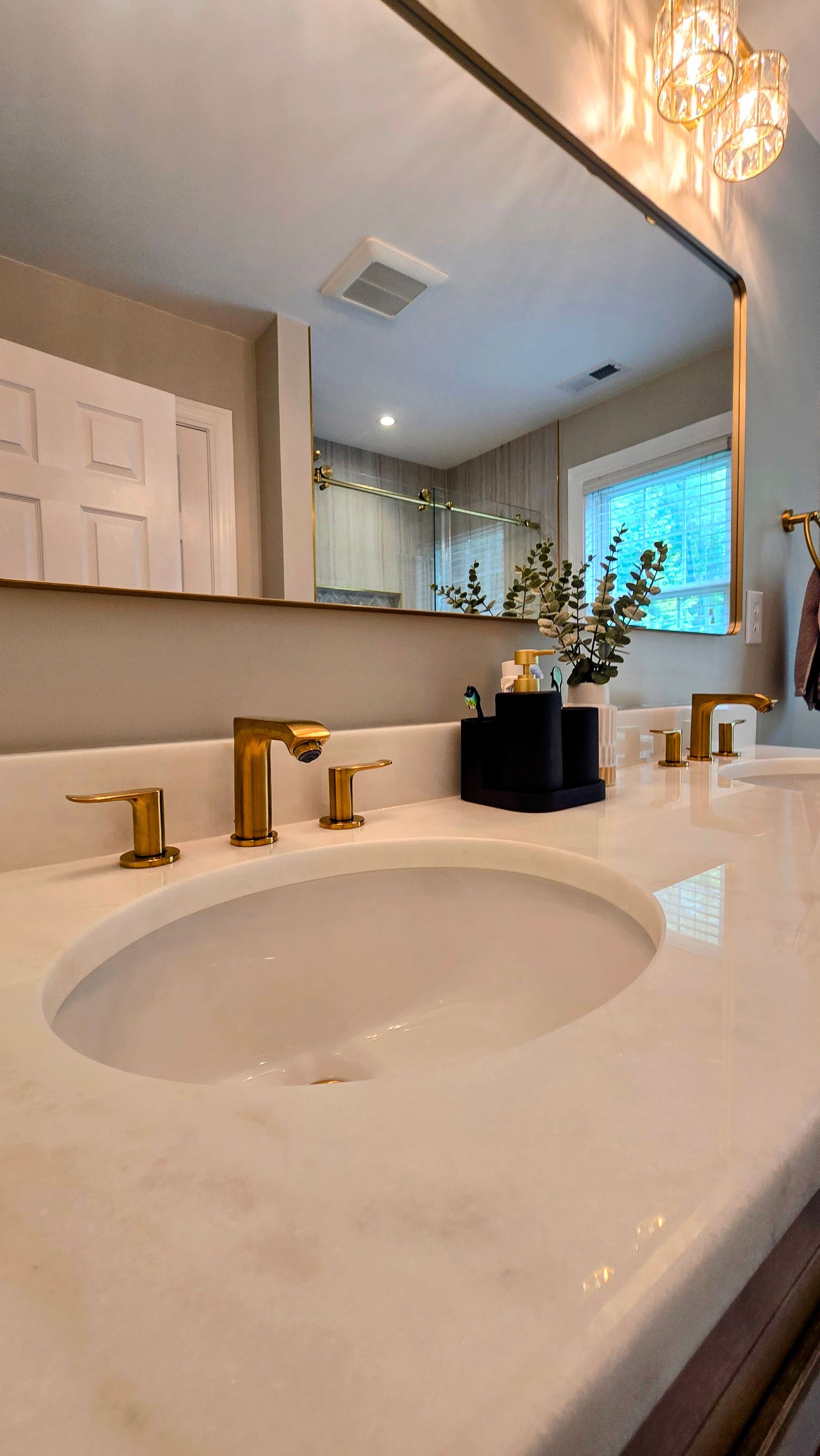 Bathroom vanity in Cheshire, CT with a white marble countertop, gold faucet, and a large mirror reflecting a window and light fixture.