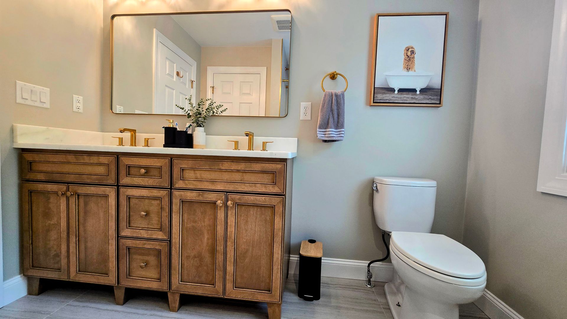 A bathroom with a wooden vanity in Cheshire, CT, a wall mirror, a toilet, and a framed picture of a bathtub hanging on the wall.